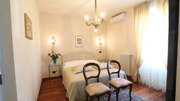Un dormitorio con una cama, dos sillas, una mesita de noche con un florero y una lámpara de pared, además de una ventana con cortinas.