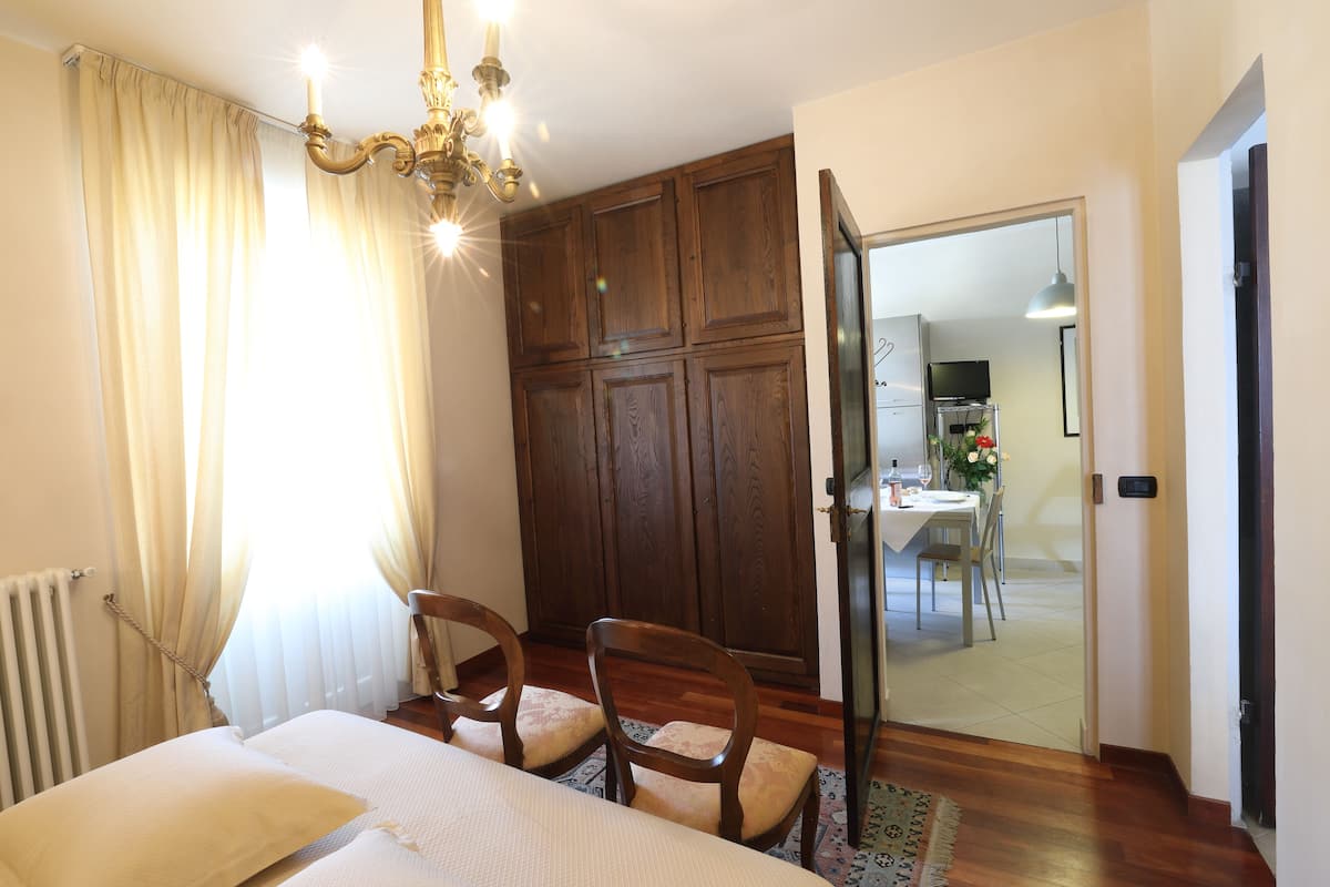 A bedroom with a bed, a wooden wardrobe, a chandelier, and a view into the dining area.