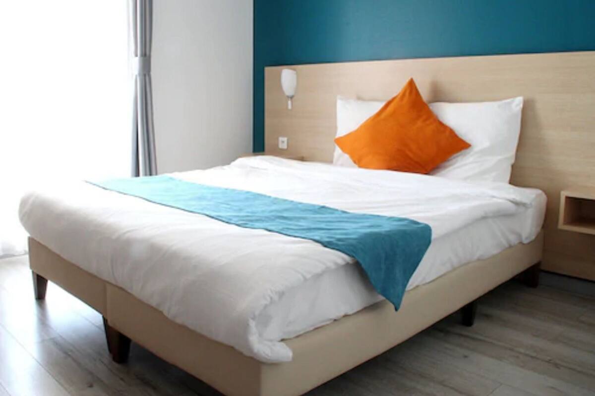 Ein ordentlich bezogenes Bett mit weißer und blauer Bettwäsche, einem orangefarbenen Kissen und einem hölzernen Kopfteil.
