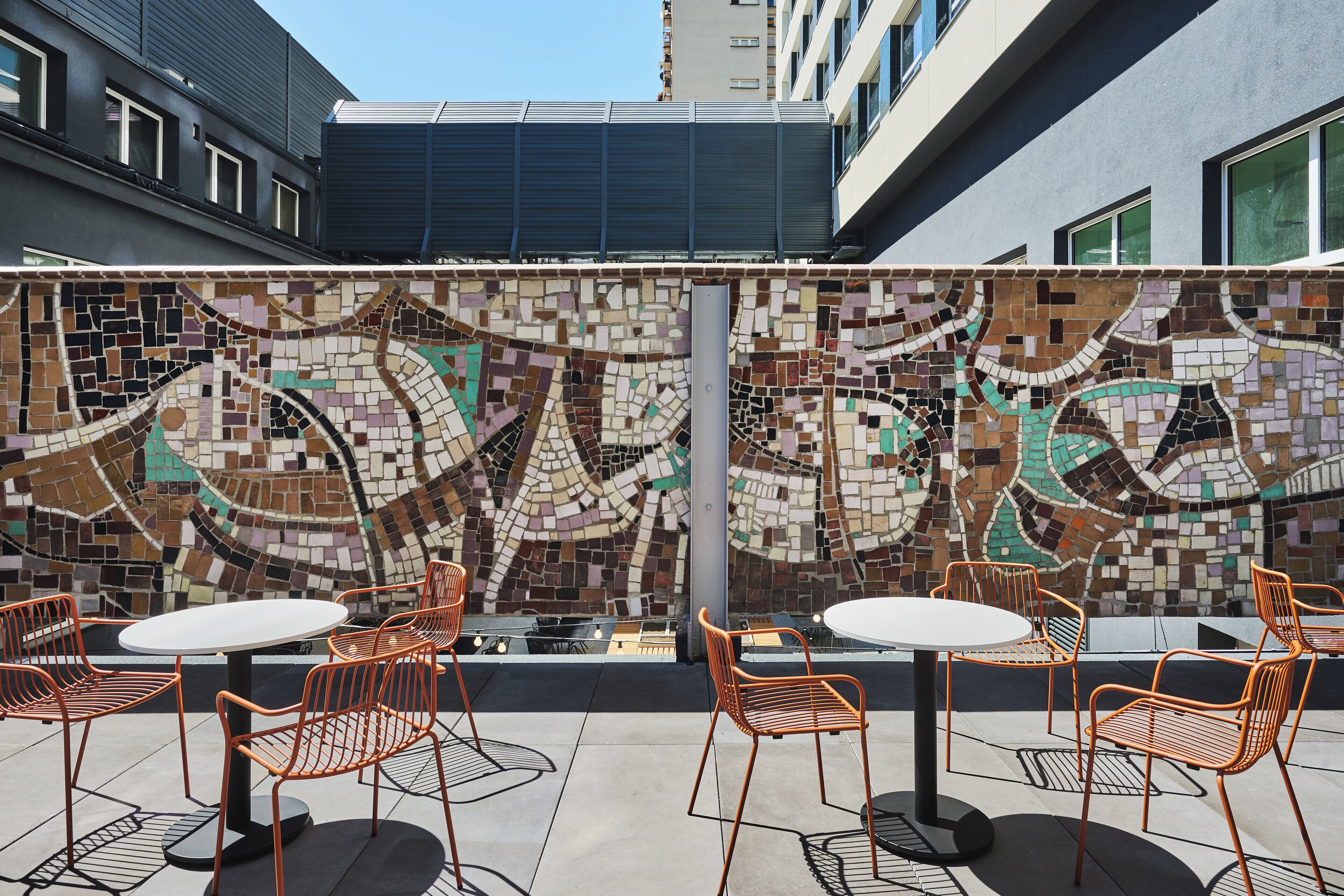 An outdoor seating area with mosaic wall art, metal chairs, and tables.