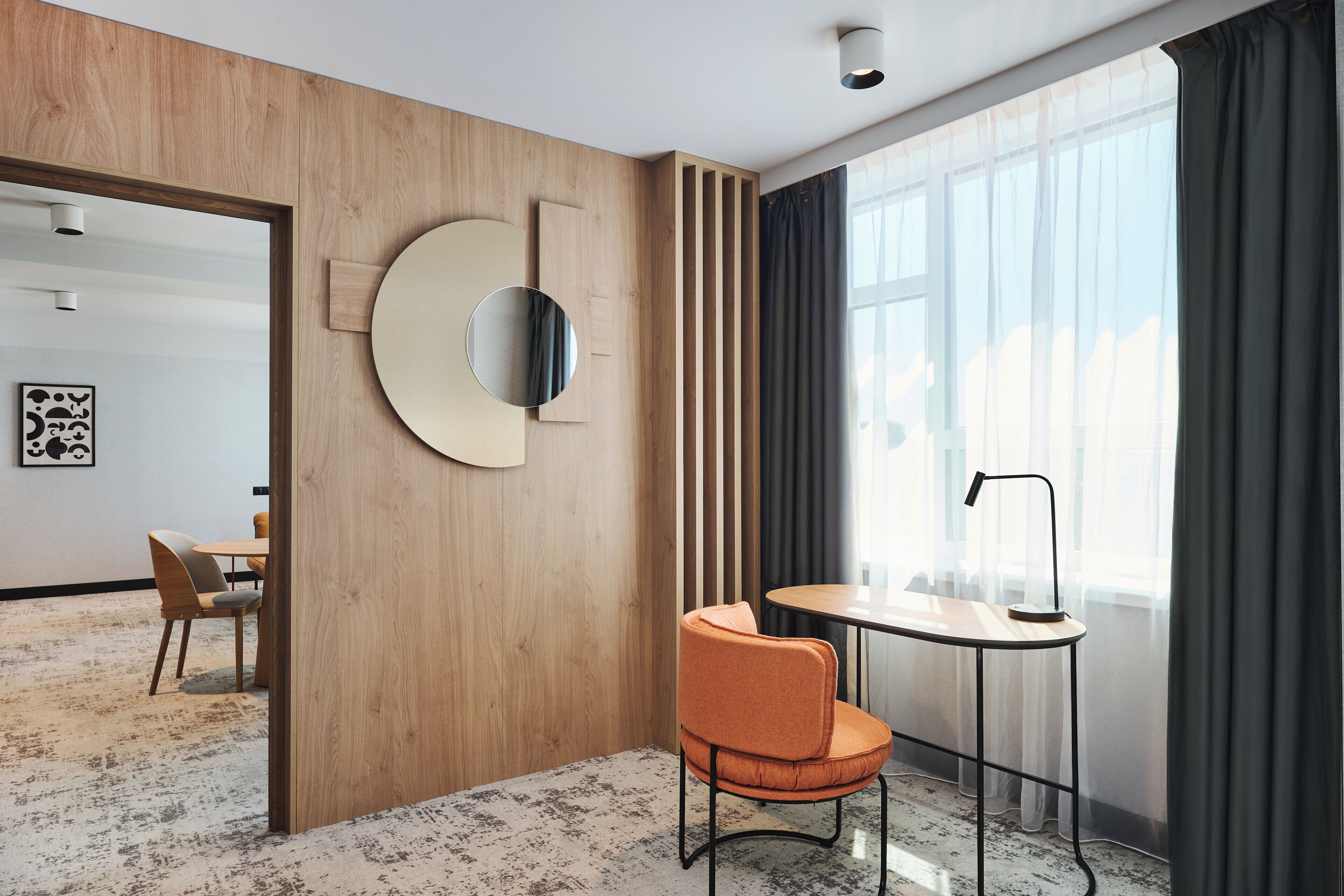 A modern hotel room with a wooden panel wall, a round mirror, a small round table with a lamp, and a view through large windows.