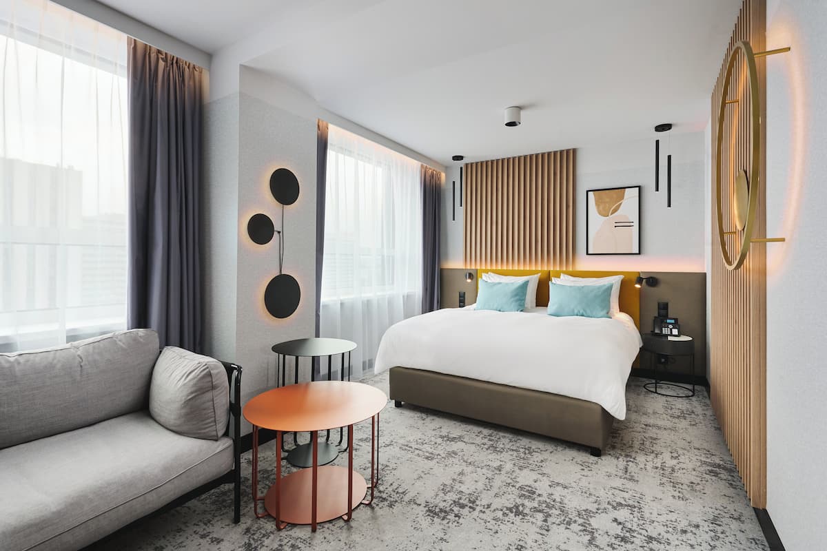Habitación de hotel moderna con una cama grande, un sofá, una mesita redonda y vistas a la ciudad a través de amplios ventanales.