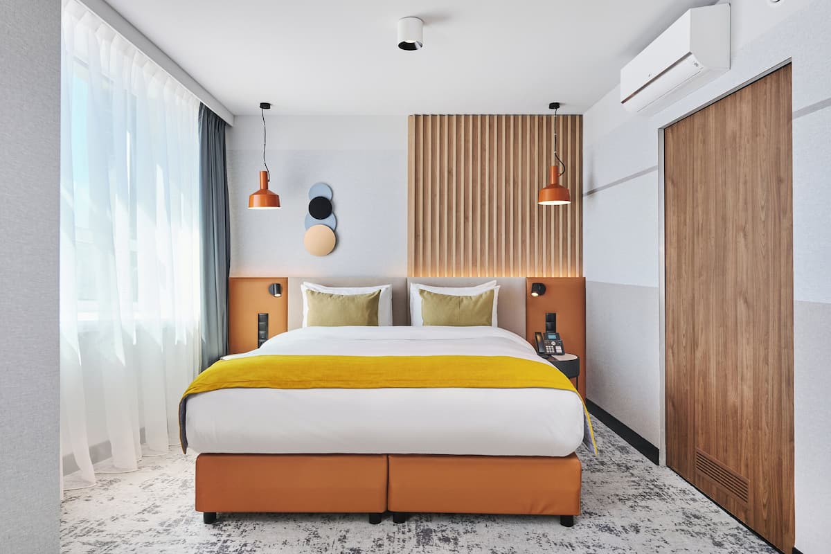Habitación de hotel moderna con una cama grande, cabecera de madera, mesita de noche con teléfono y una ventana amplia con cortinas.