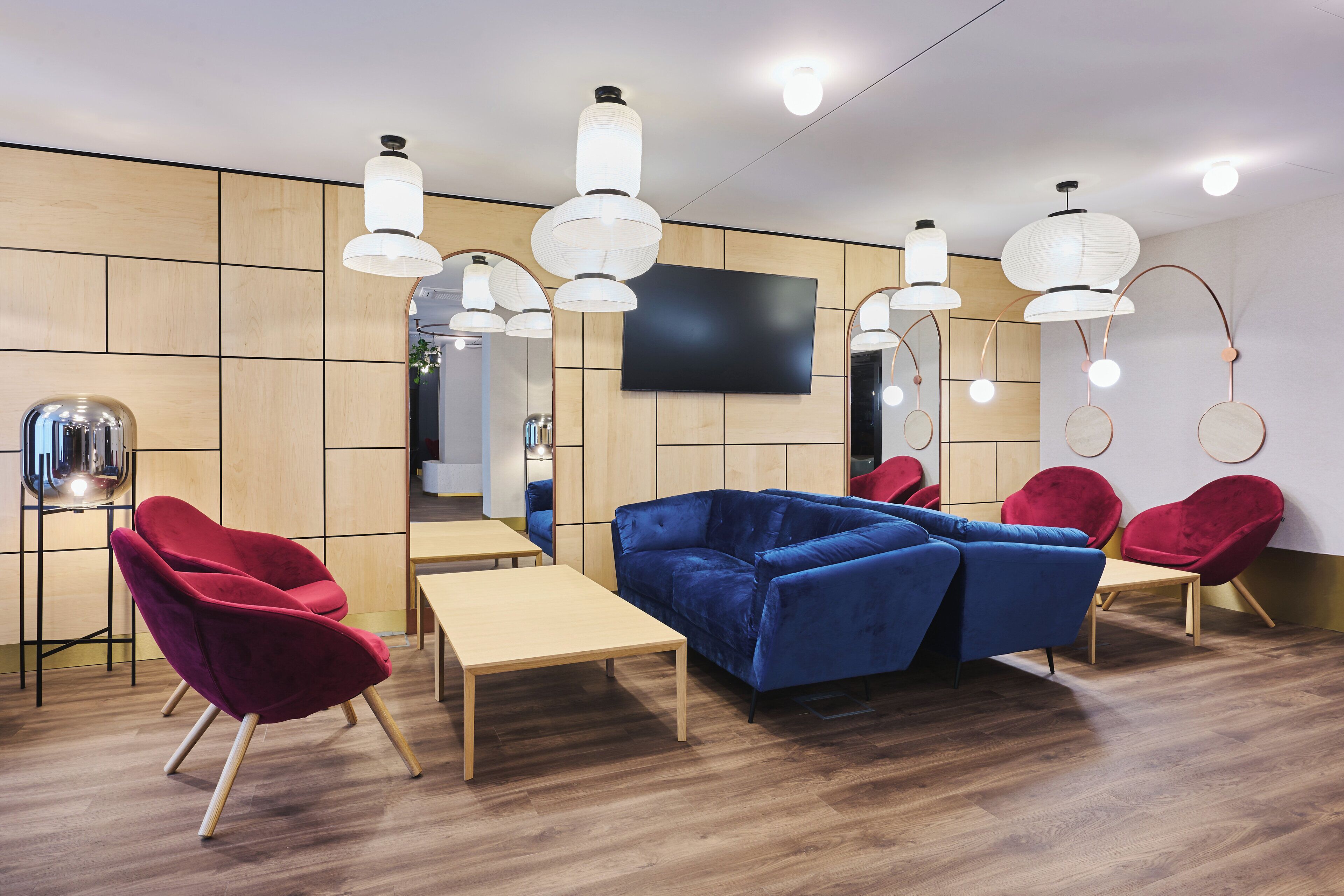 A modern interior with a blue sofa, red chairs, wooden tables, and hanging pendant lights.