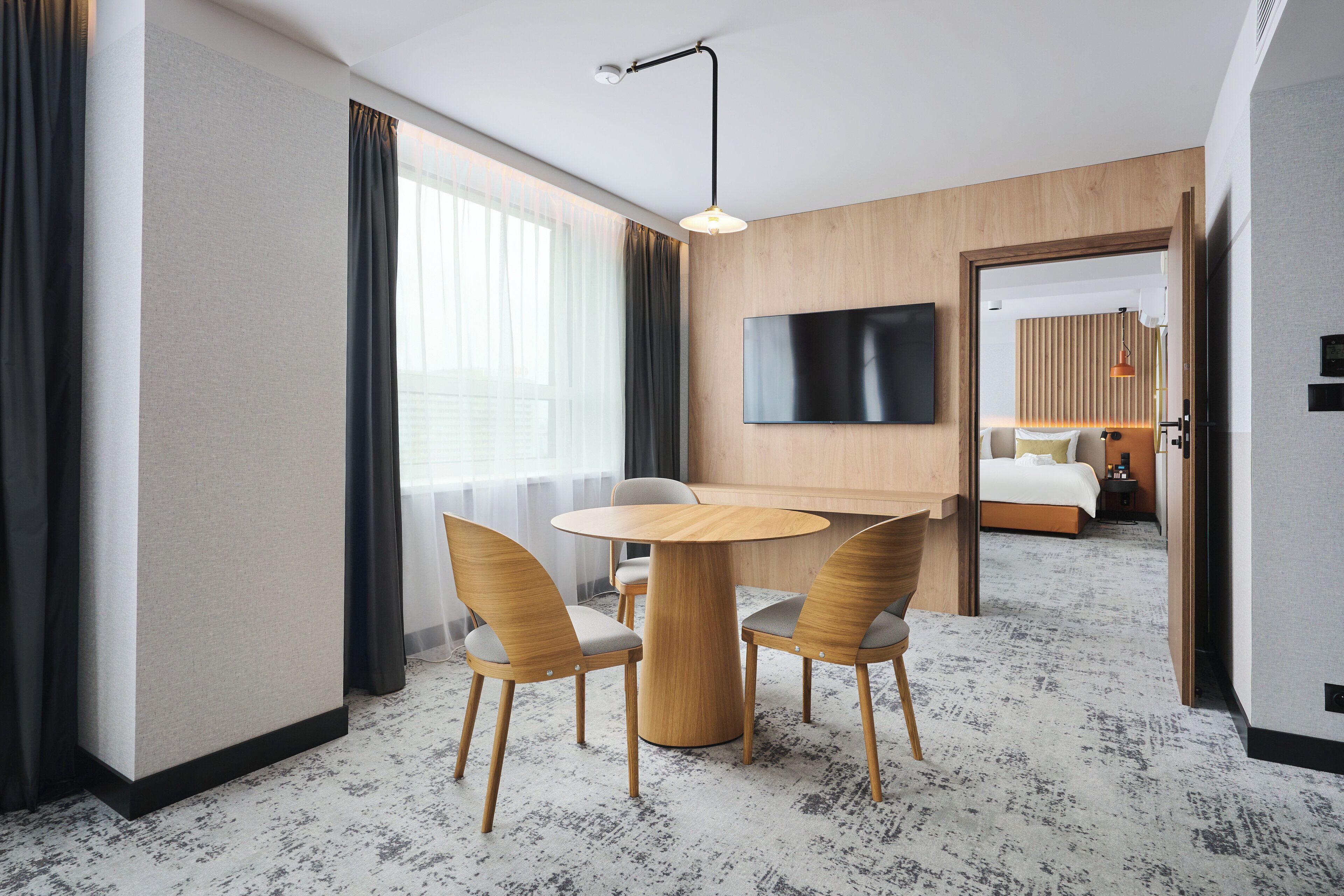 A hotel room with a round wooden table, two chairs, a flat-screen TV mounted on the wall, and a bed with a wooden headboard.