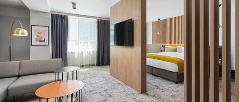 A modern hotel room with a sofa, a small table, a bed with a yellow blanket, a TV mounted on a wooden panel, and a large window with curtains.