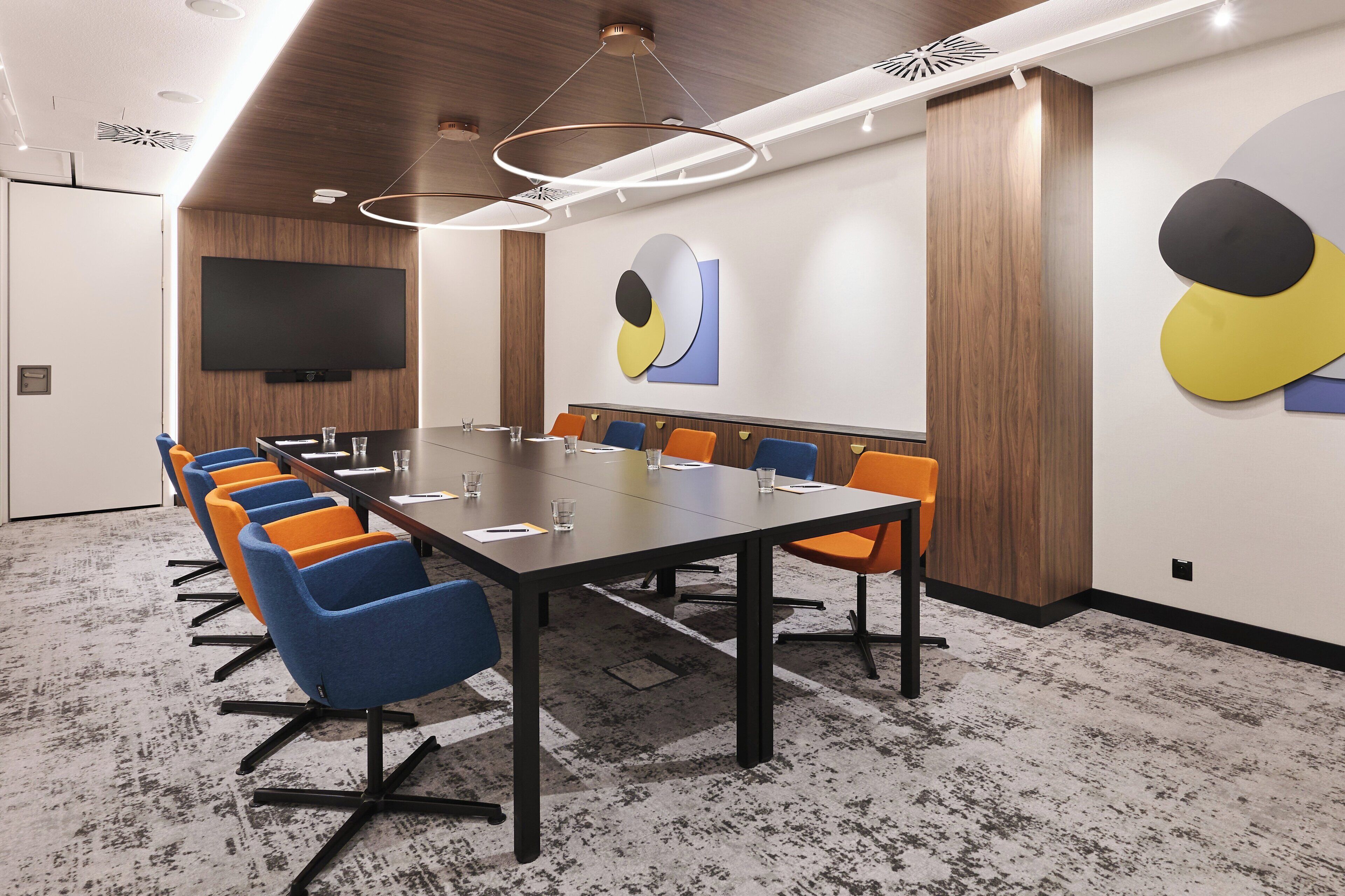 A conference room with a long table, blue and orange chairs, a flat-screen TV, and abstract wall art.