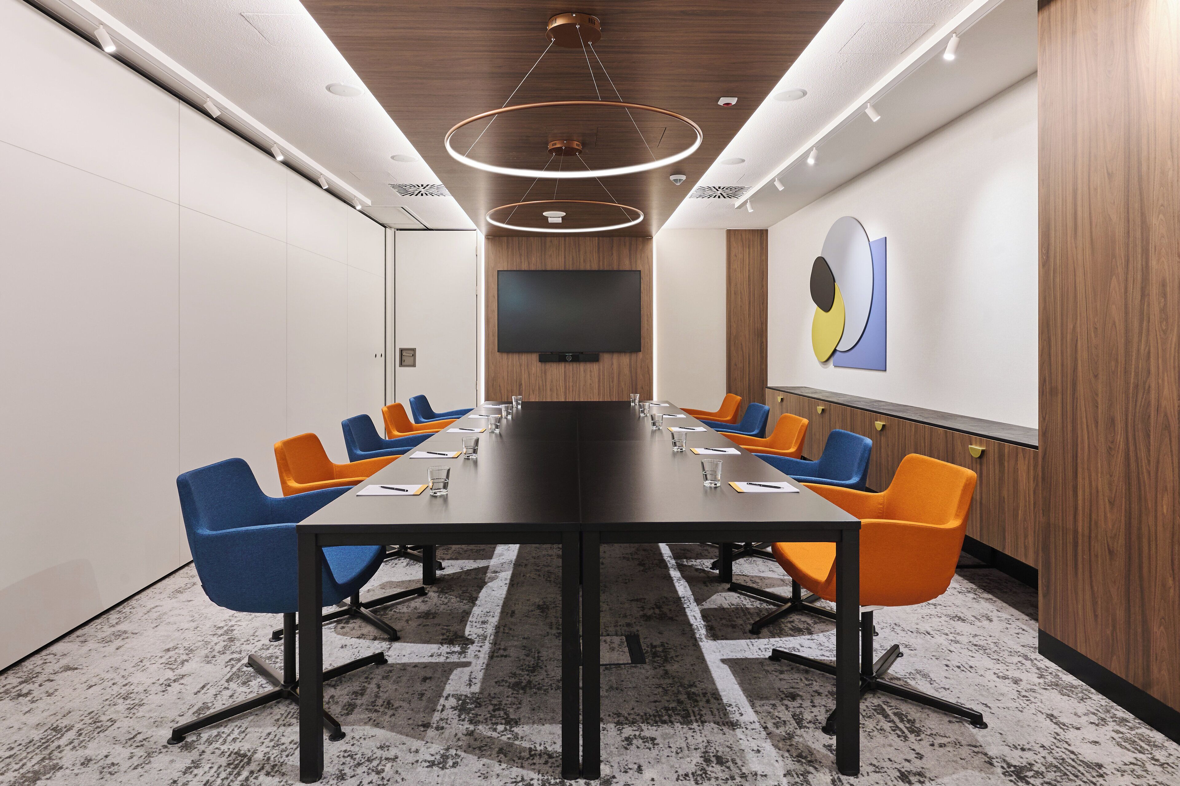 Een vergaderruimte met een lange tafel, blauwe en oranje stoelen, een flatscreen televisie en een houten accentmuur.