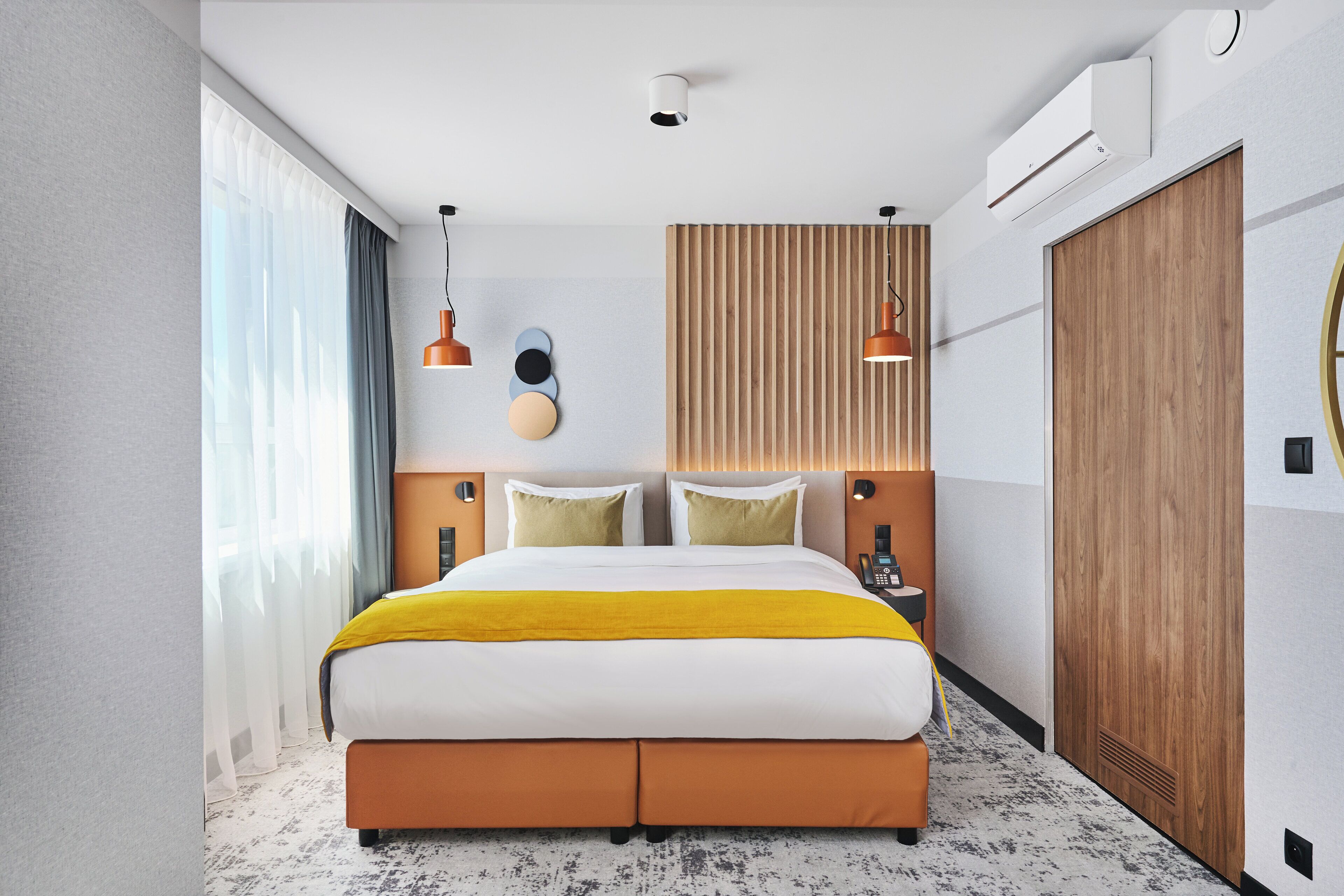 A modern hotel room with a large bed, wooden headboard, two bedside tables, a built-in wardrobe, and a large window with curtains.