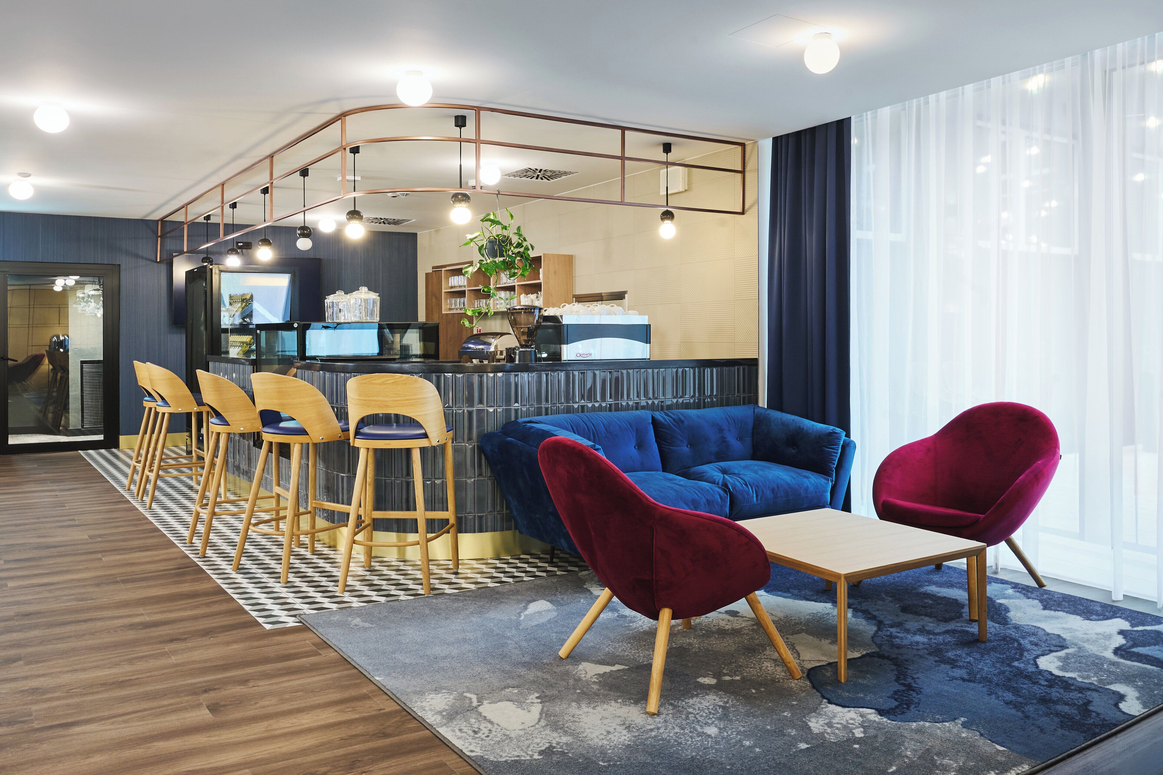 Un área de descanso moderna con un sofá azul, sillas rojas, taburetes de madera y una estación de café.