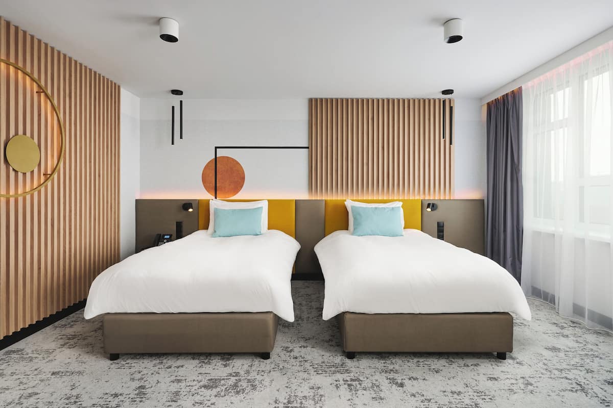 Una habitación de hotel moderna con dos camas, una pared de acento de madera, un ventanal con cortinas y una lámpara circular.