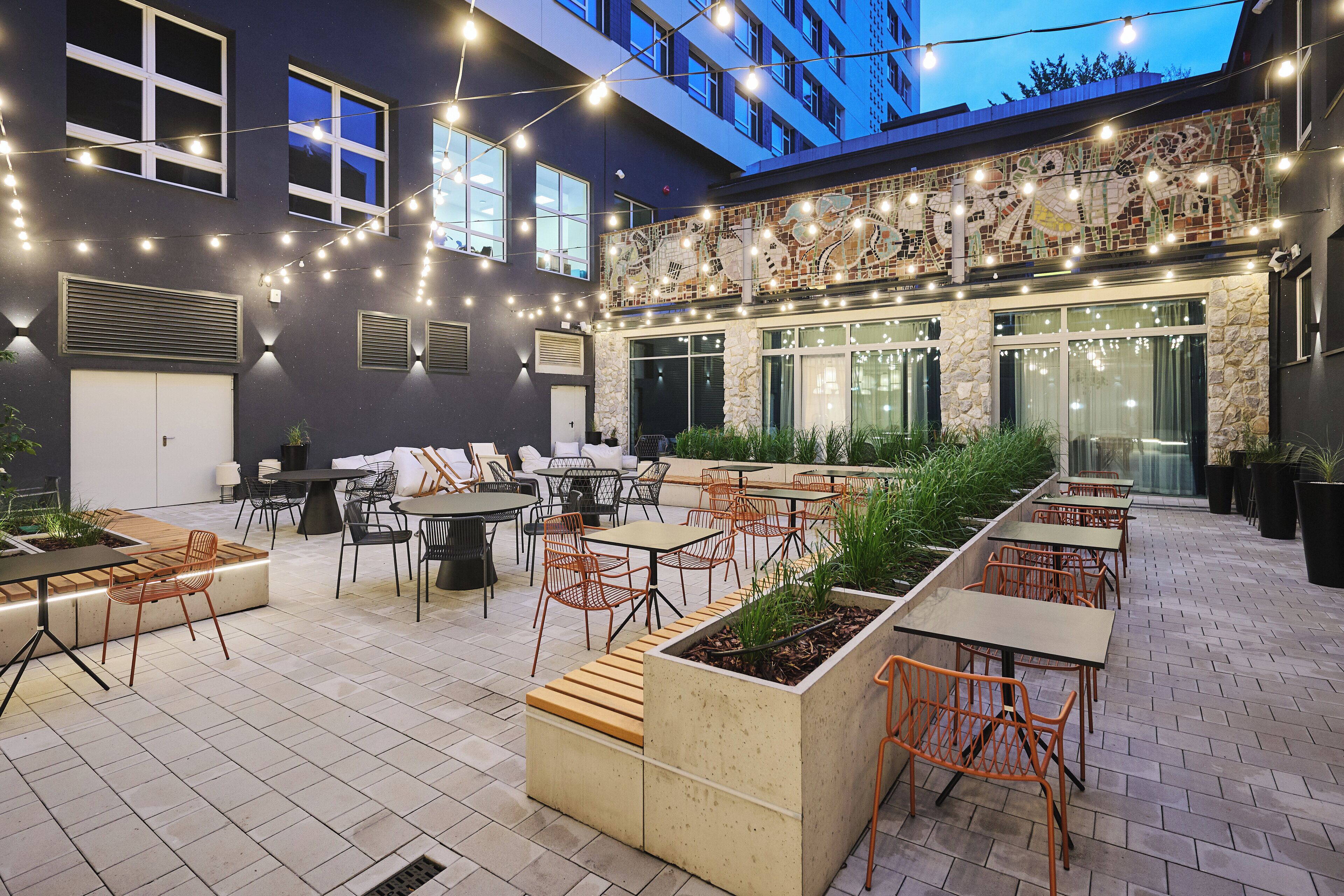 An outdoor patio with tables and chairs, string lights, and a mosaic wall.