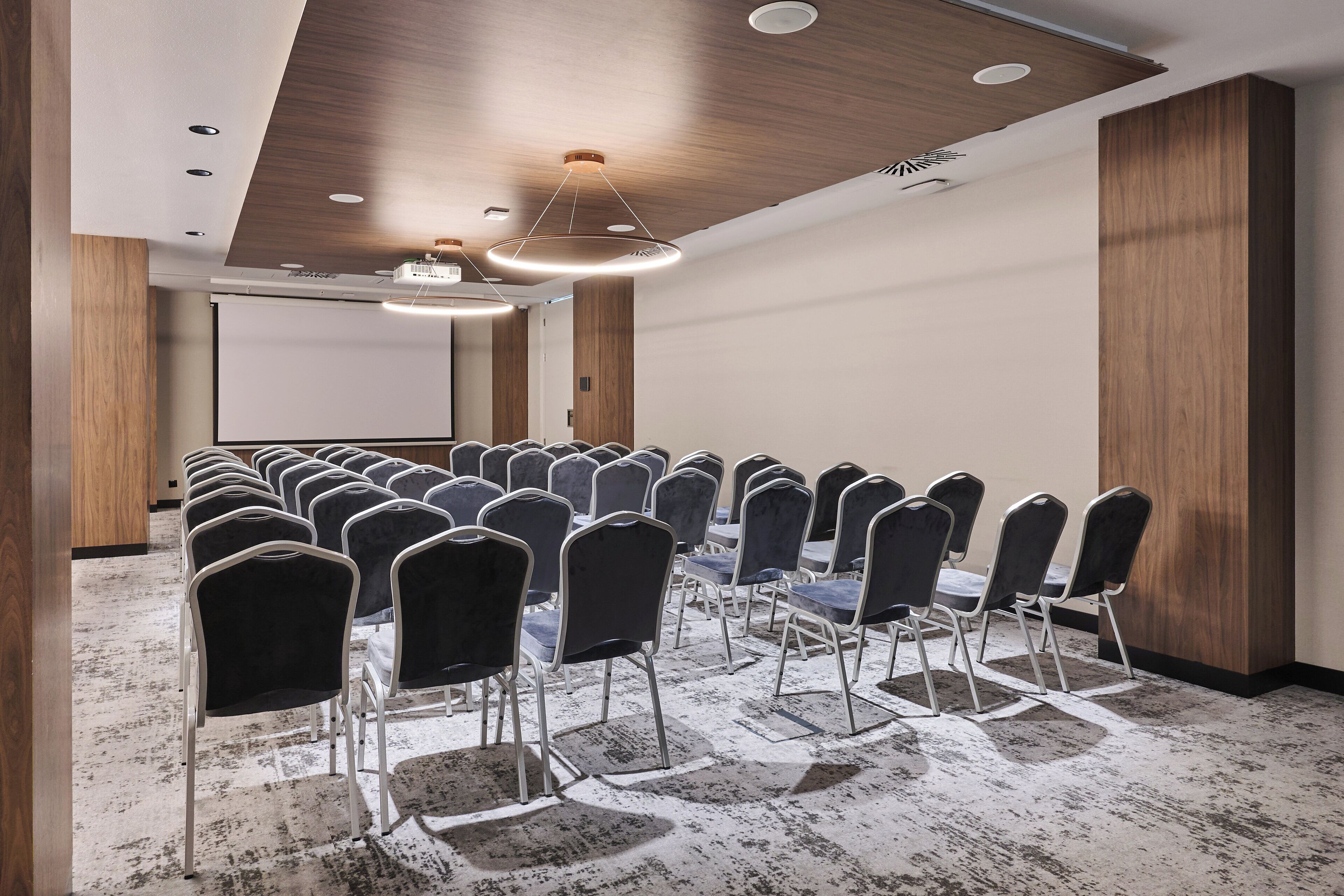 A conference room with rows of chairs and tables, wooden paneling on the walls, and a projector screen.