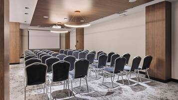 Une salle de conférence avec des rangées de chaises et de tables, des panneaux de bois sur les murs et un écran de projection.