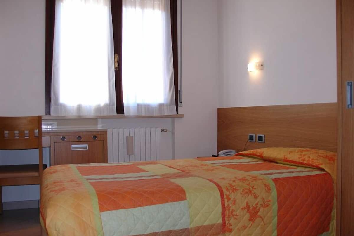 A bedroom with a wooden bed, a desk with a chair, a radiator, and a window with curtains.
