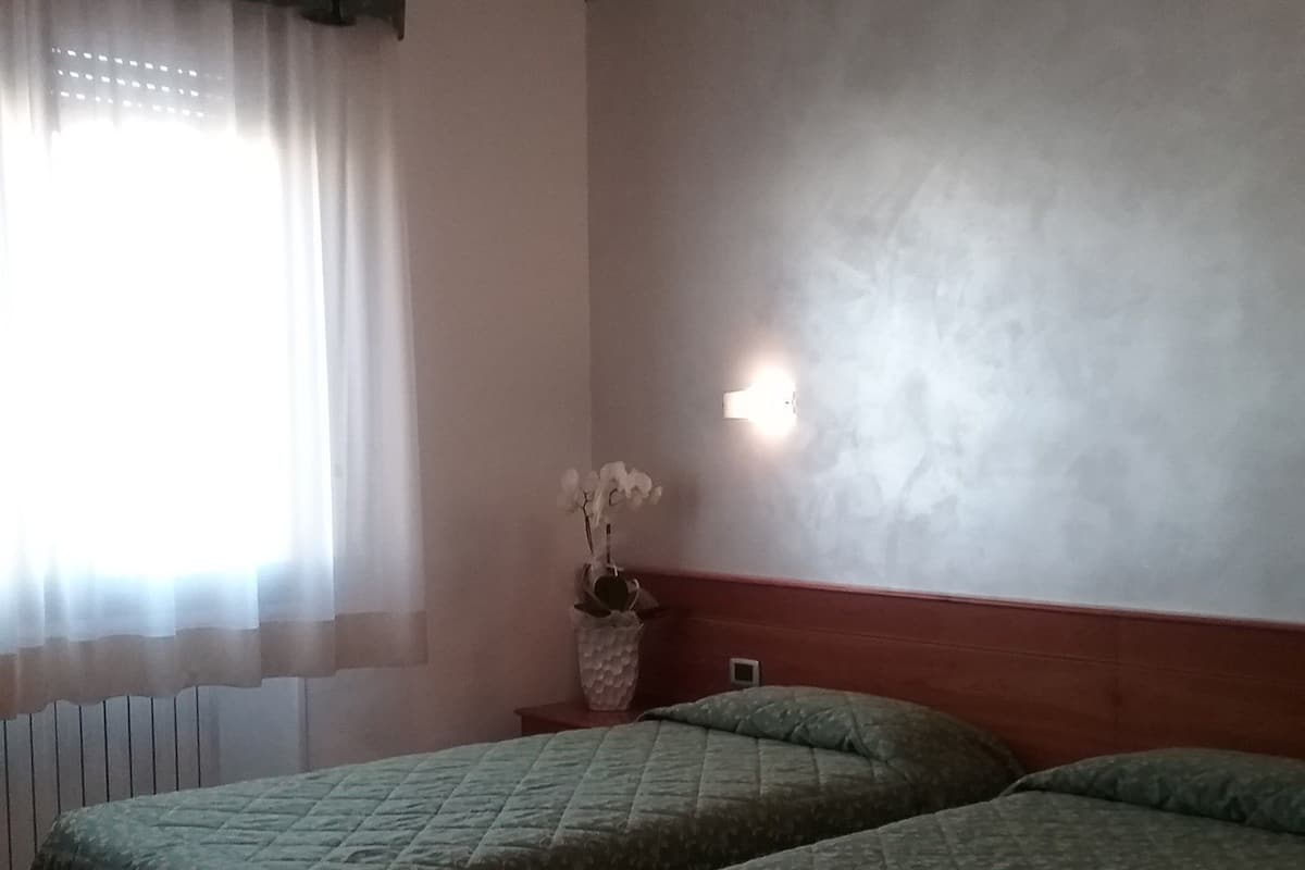 A room with two beds, a window with sheer curtains, a wall-mounted light, and a radiator.