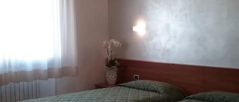 A room with two beds, a window with sheer curtains, a wall-mounted light, and a radiator.