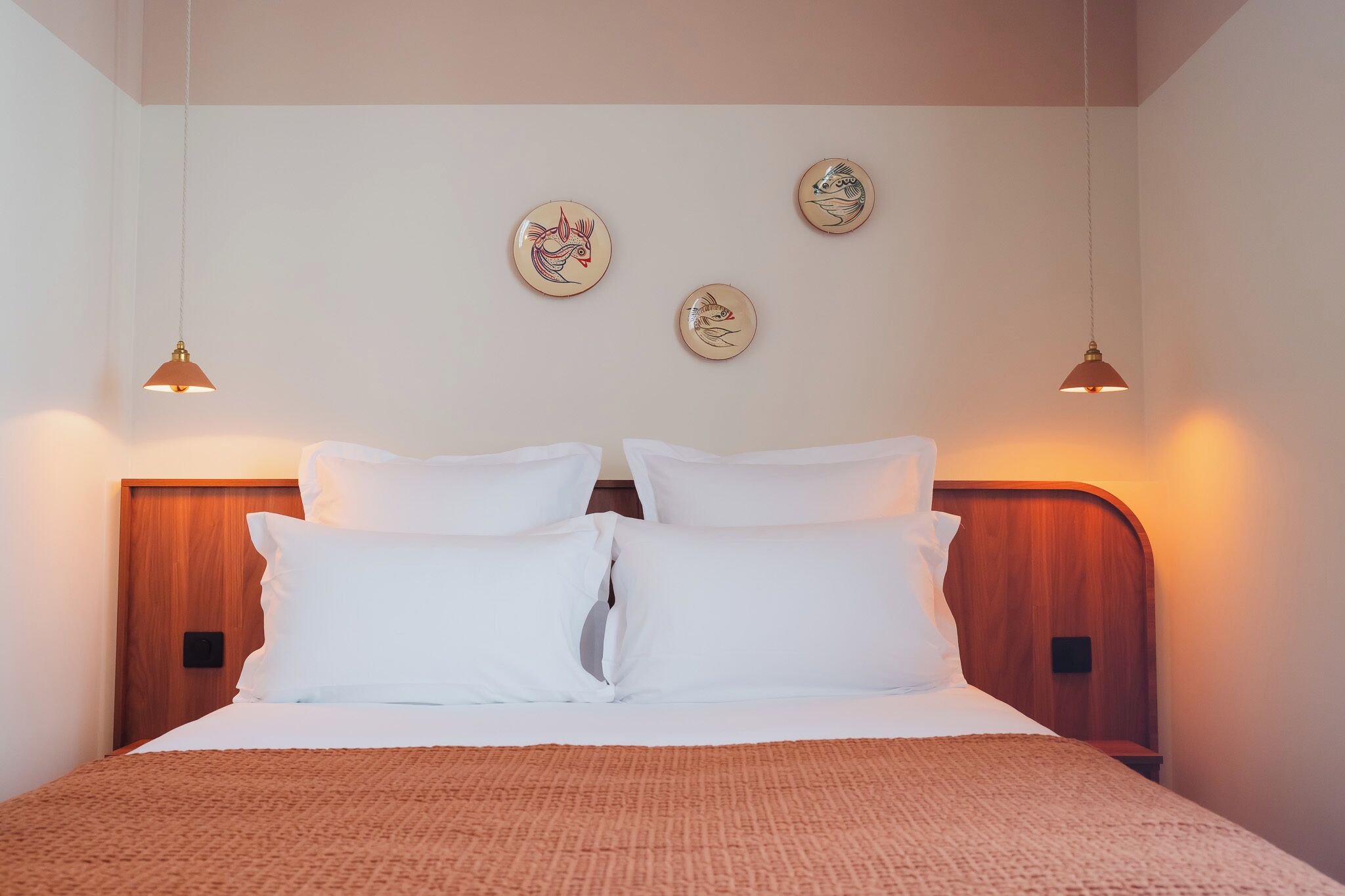 A neatly made bed with white linens and a brown throw, a wooden headboard, and three wall-mounted light fixtures with decorative plates.