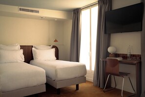A hotel room with two beds, a desk with a chair, a television, and a window with curtains. - Lala Hotel (Paris)