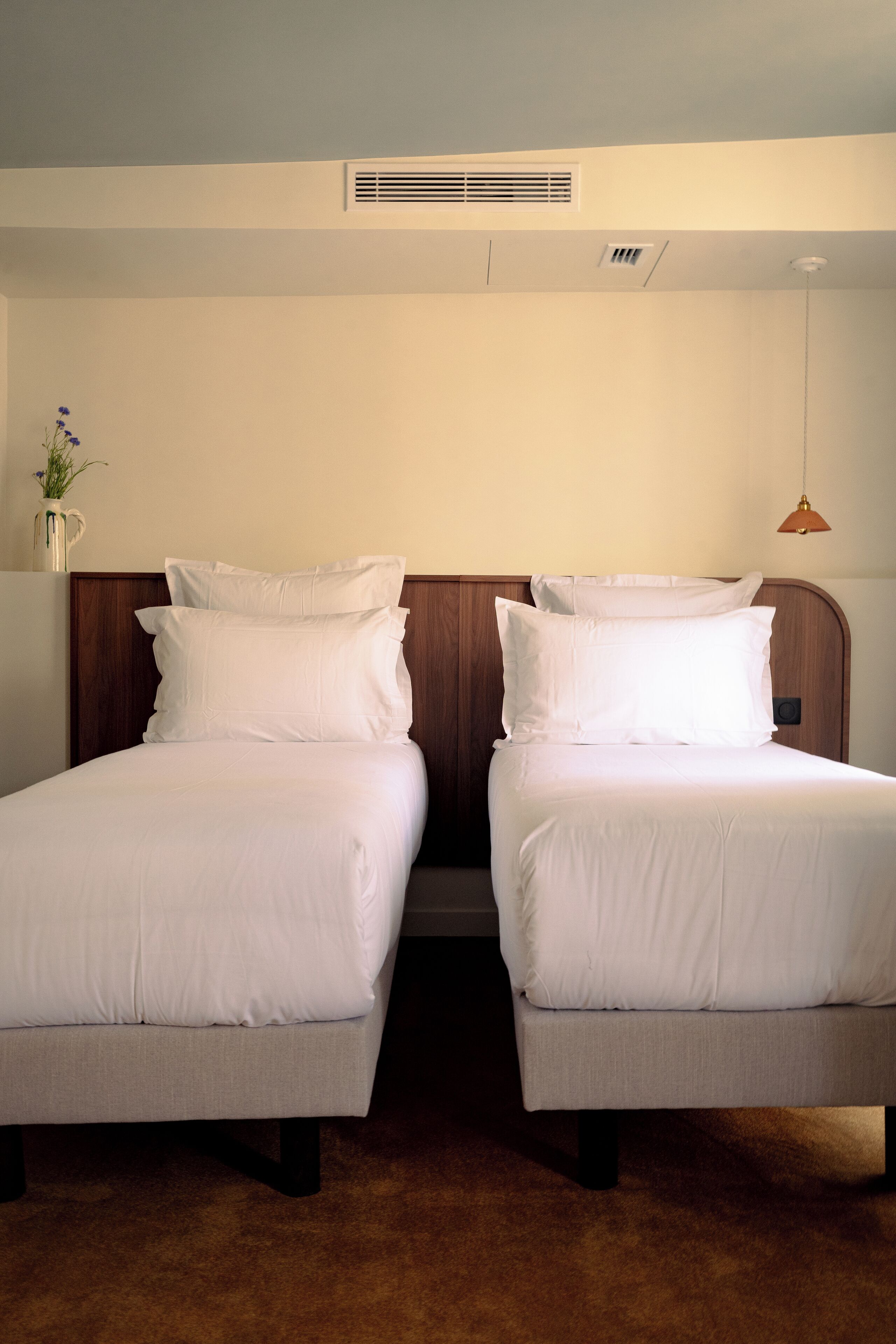 Una habitación de hotel con dos camas individuales, un cabecero de madera, un pequeño jarrón con flores y una lámpara colgante.