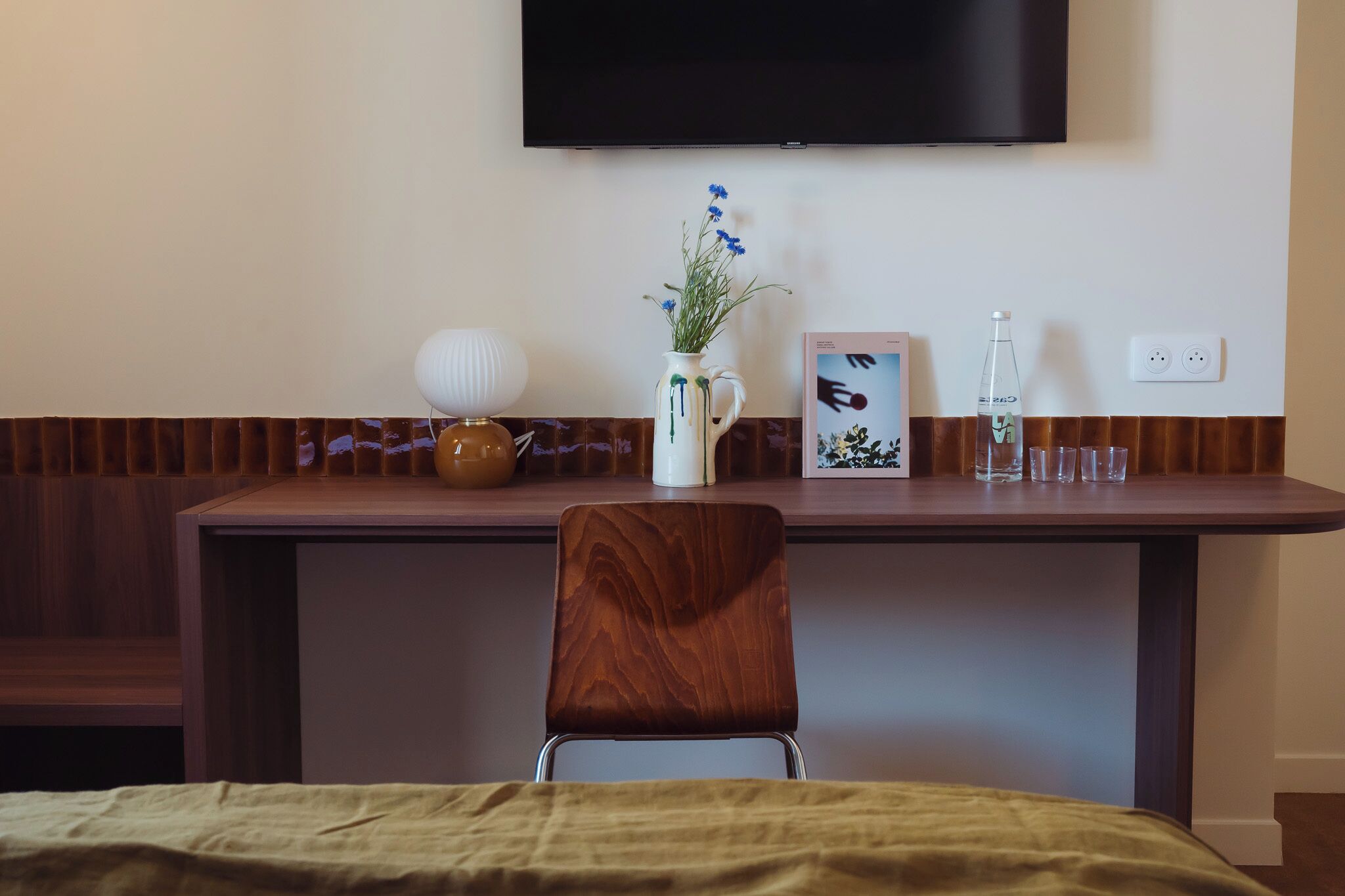 Una camera d'albergo con una scrivania in legno, un vaso bianco con fiori blu, un quadro incorniciato, una bottiglia e un bicchiere.
