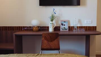 Una camera d'albergo con una scrivania in legno, un vaso bianco con fiori blu, un quadro incorniciato, una bottiglia e un bicchiere.