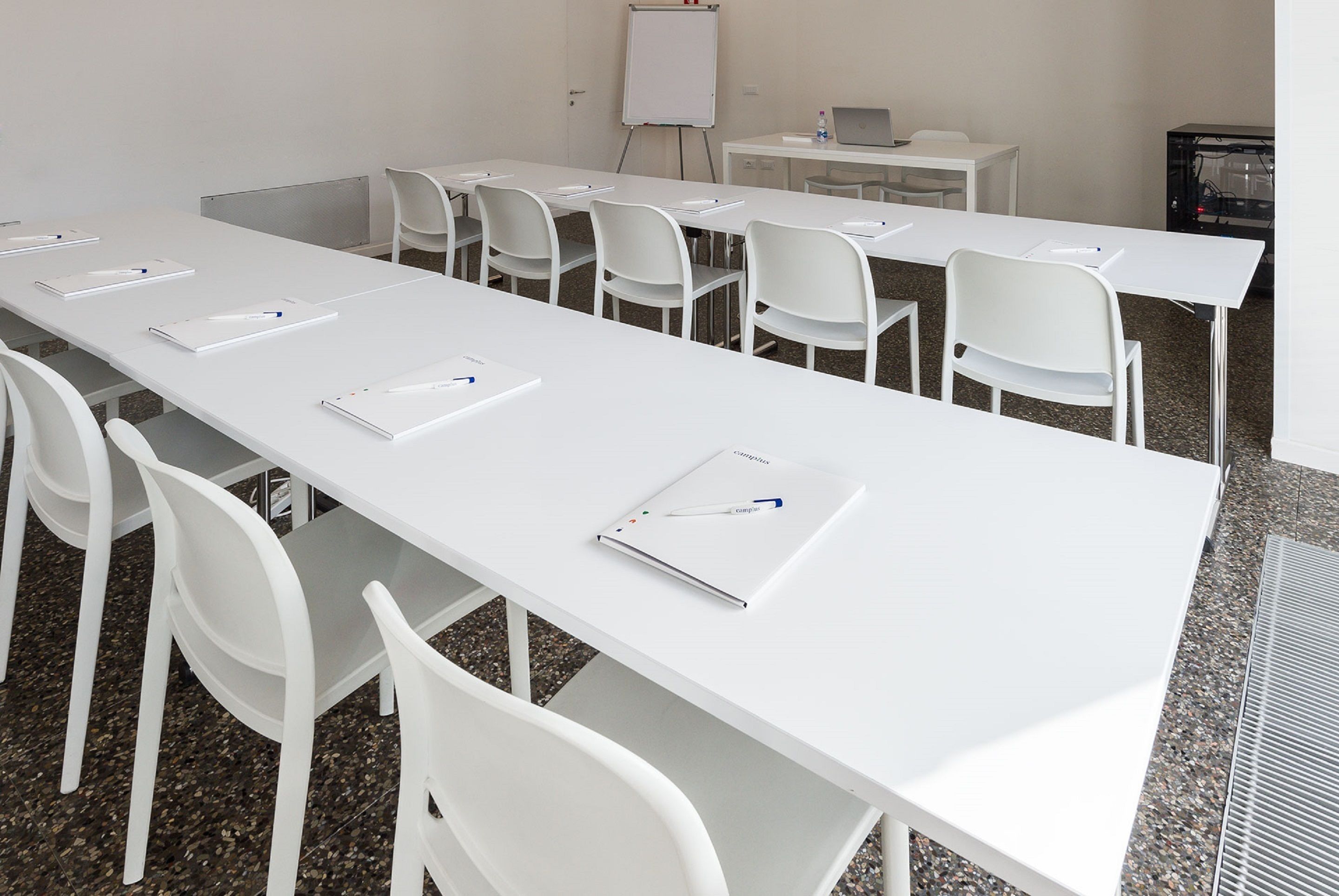 Une salle de conférence avec une longue table blanche, des chaises blanches, un tableau blanc et un ordinateur portable posé sur une petite table.