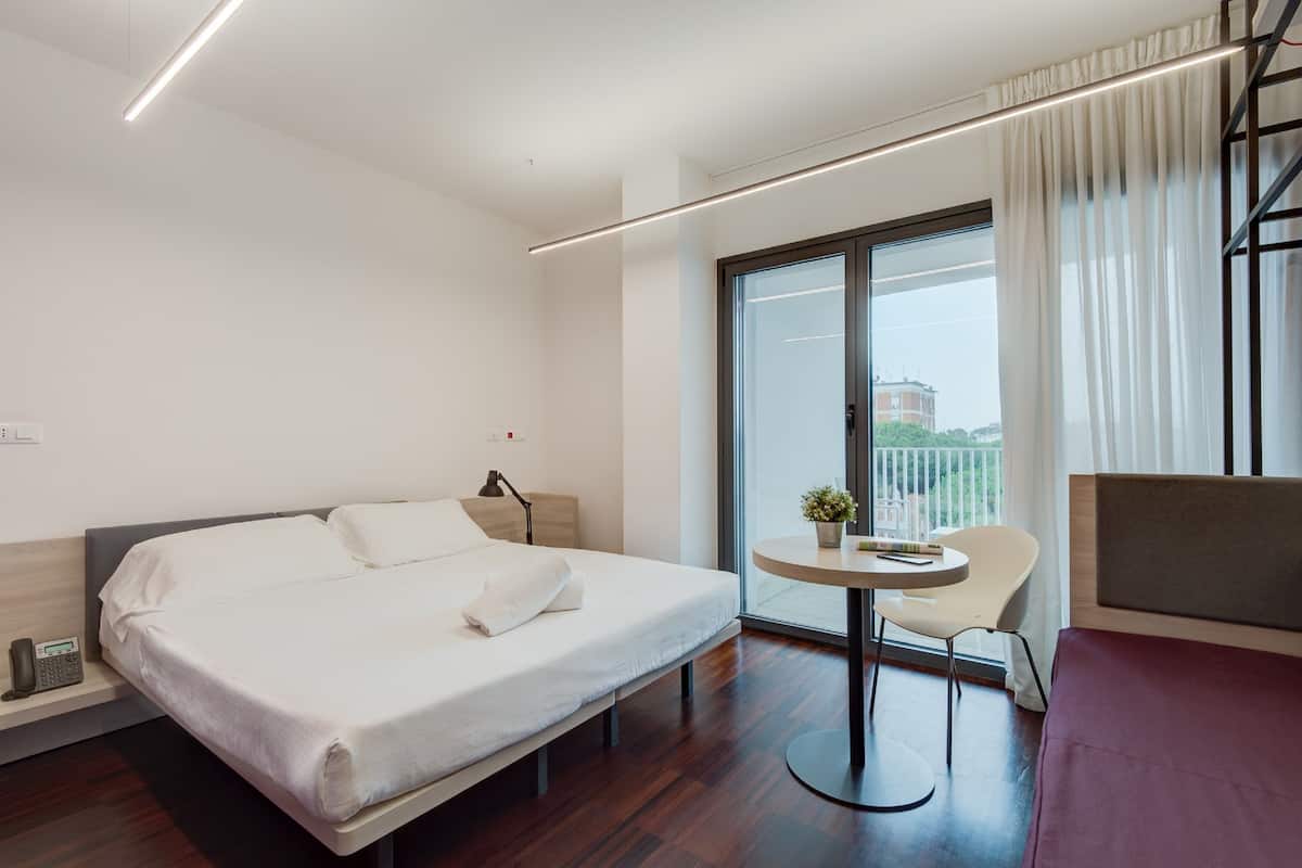 Habitación de hotel moderna con una cama grande, una mesita redonda pequeña con una silla, una lámpara de noche y vistas a la ciudad a través de un ventanal amplio.