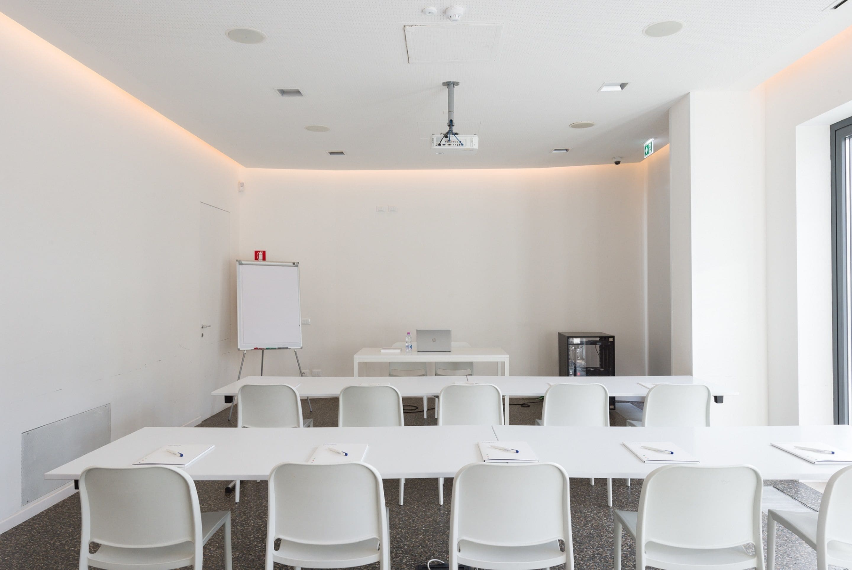 A conference room with white tables and chairs, a whiteboard, a projector, and a laptop on a table.