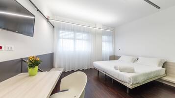 A modern hotel room with a bed, a desk, a chair, a TV mounted on the wall, and a window with curtains.