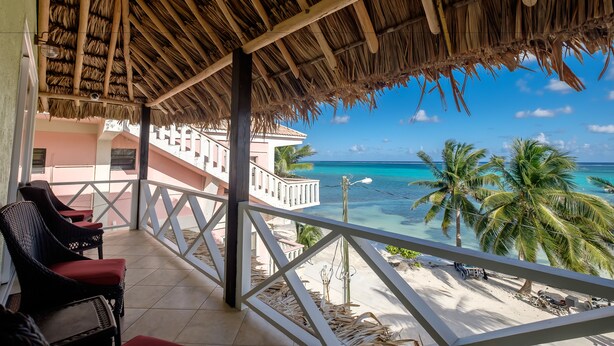 Un balcón con techo de paja, con vista a una playa con palmeras y agua turquesa.