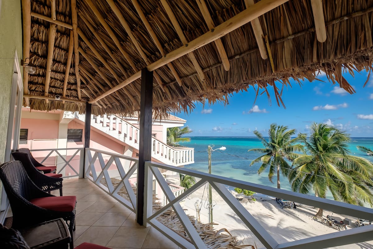 Un balcón con techo de paja, con vista a una playa con palmeras y agua turquesa.