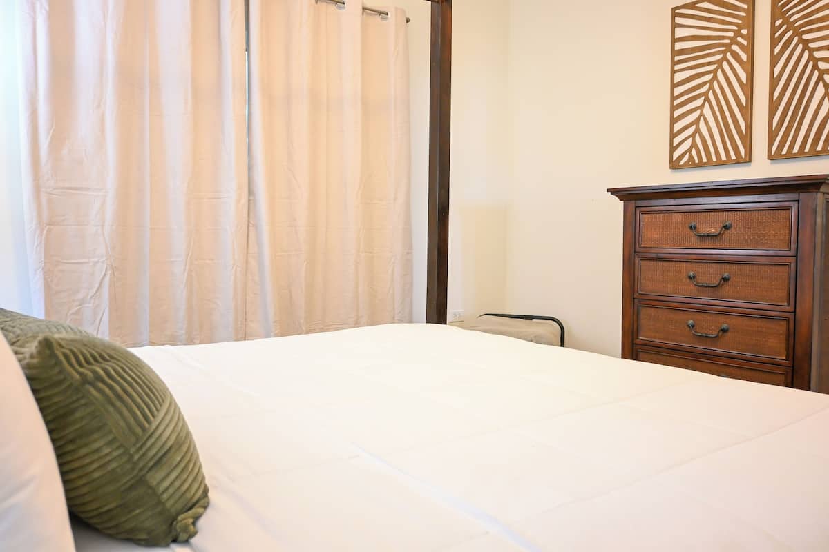 Una cama bien hecha con un almohadón verde, un aparador de madera con cajones y un panel decorativo en la pared.