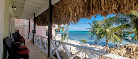 A covered balcony with wicker chairs, a thatched roof, and a view of the beach and ocean.