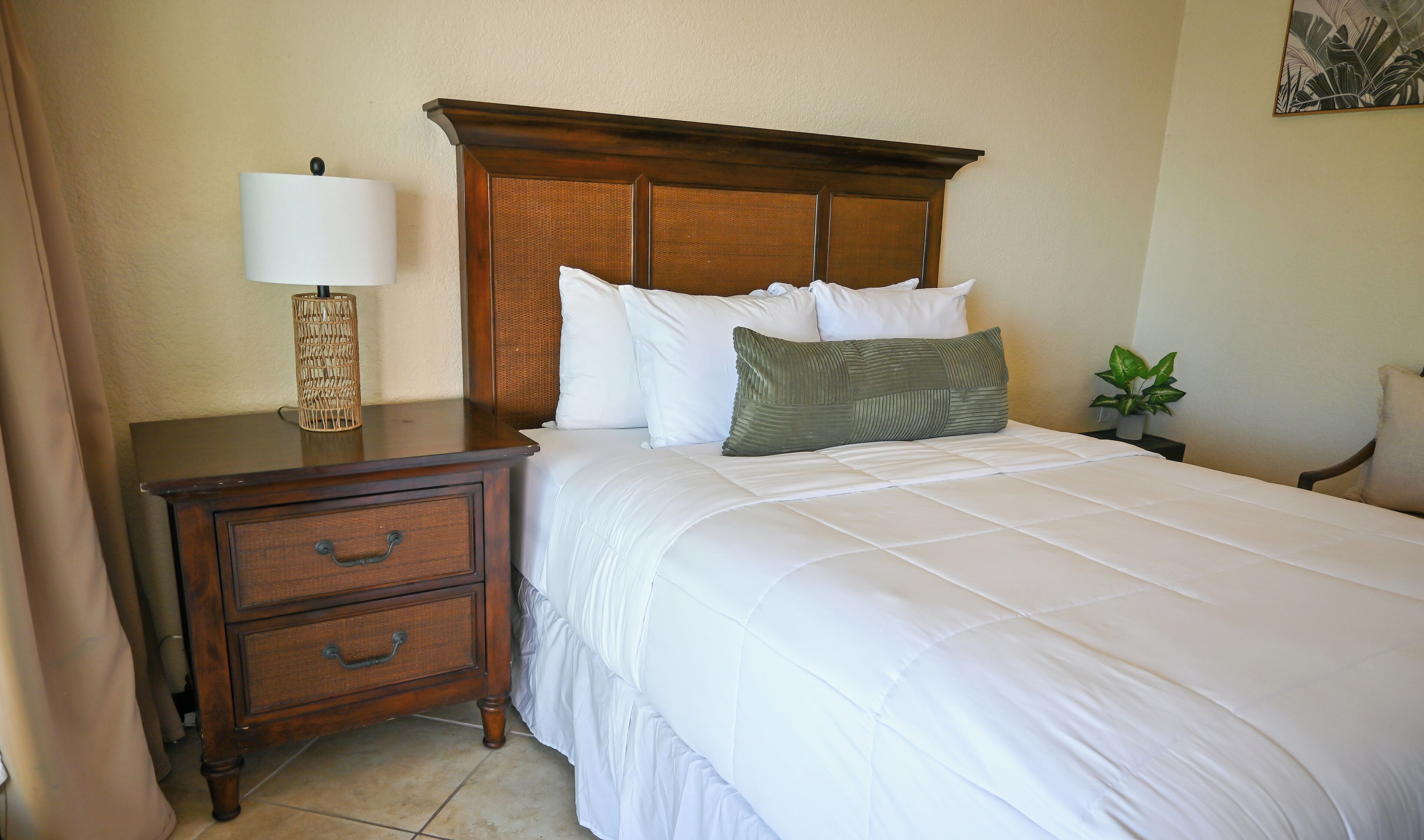 A neatly made bed with white bedding and a wooden headboard, a nightstand with a lamp and a plant, and a small drawer unit.