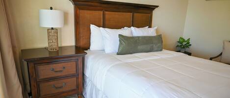 Una cama bien hecha con ropa blanca y un cabecero de madera, una mesita de noche con lámpara y planta, y un pequeño aparador.