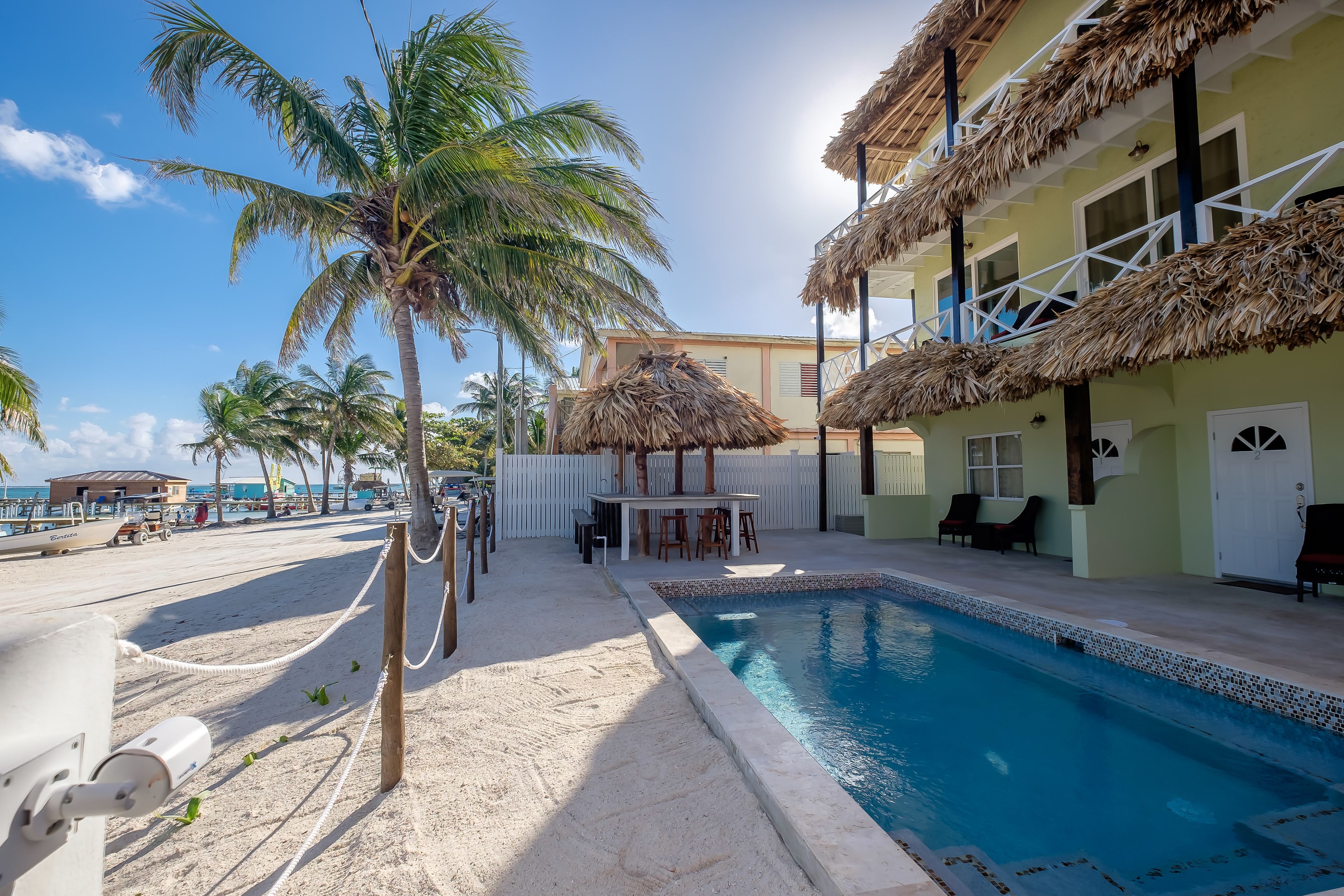 A pool area with a thatched-roof structure, palm trees, and a view of the beach.