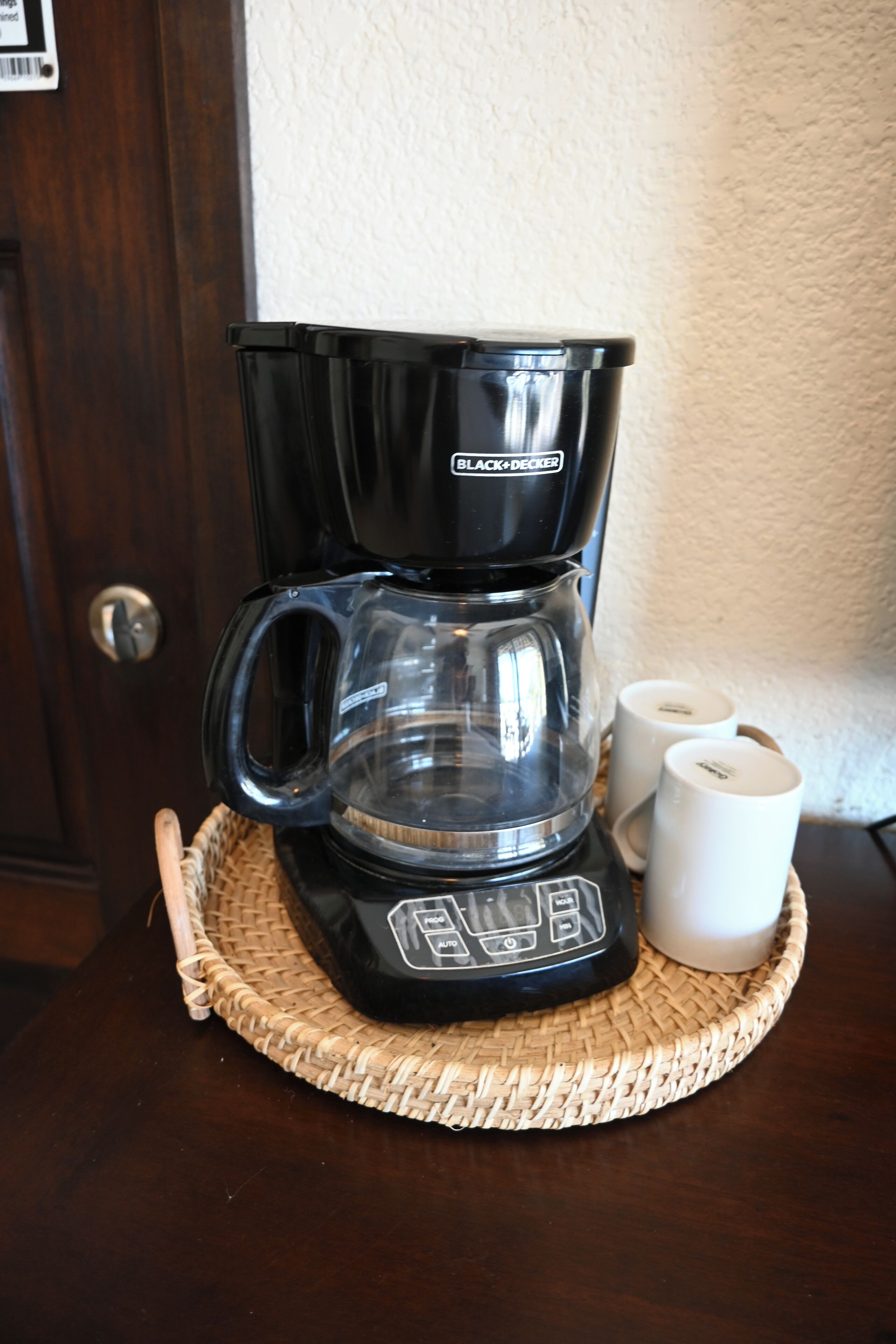 Une cafetière noire avec une verseuse en verre et deux tasses blanches posées sur un sous-verre en fibres tressées.
