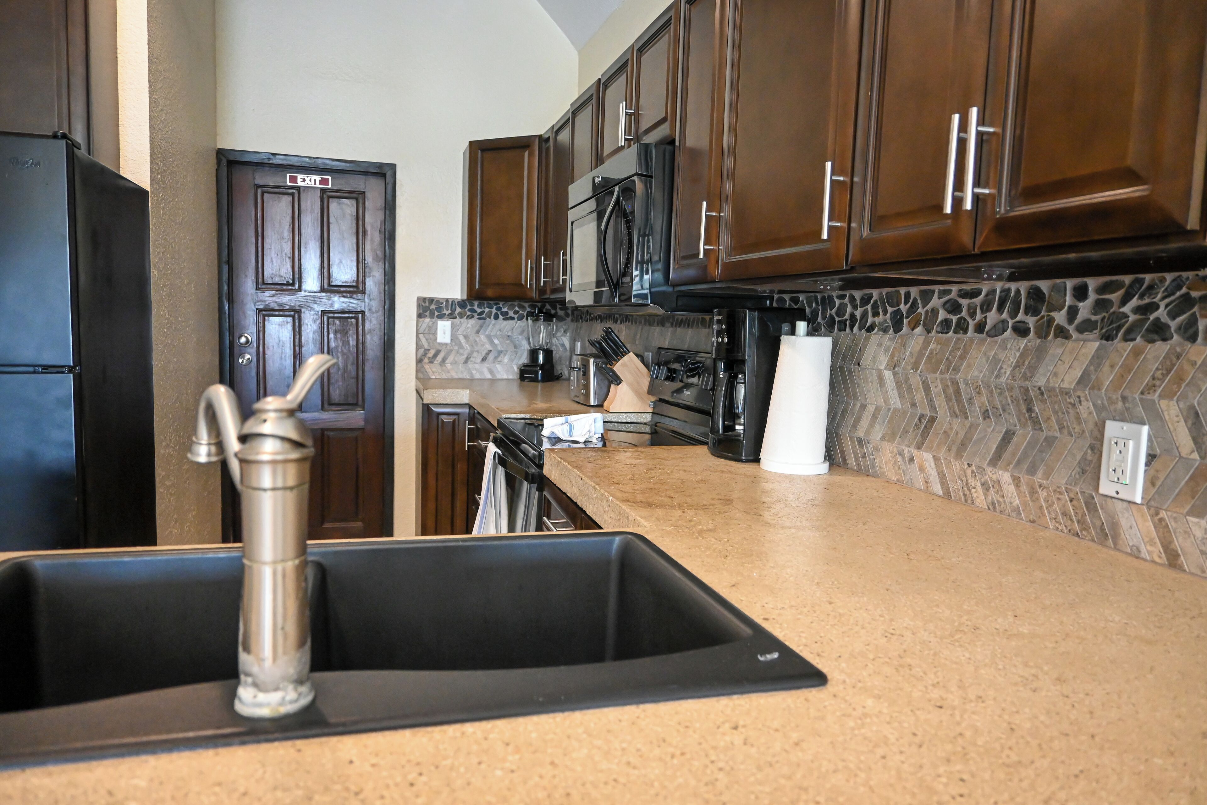 A kitchen with dark wooden cabinets, a black refrigerator, a stainless steel sink, and a stone backsplash.