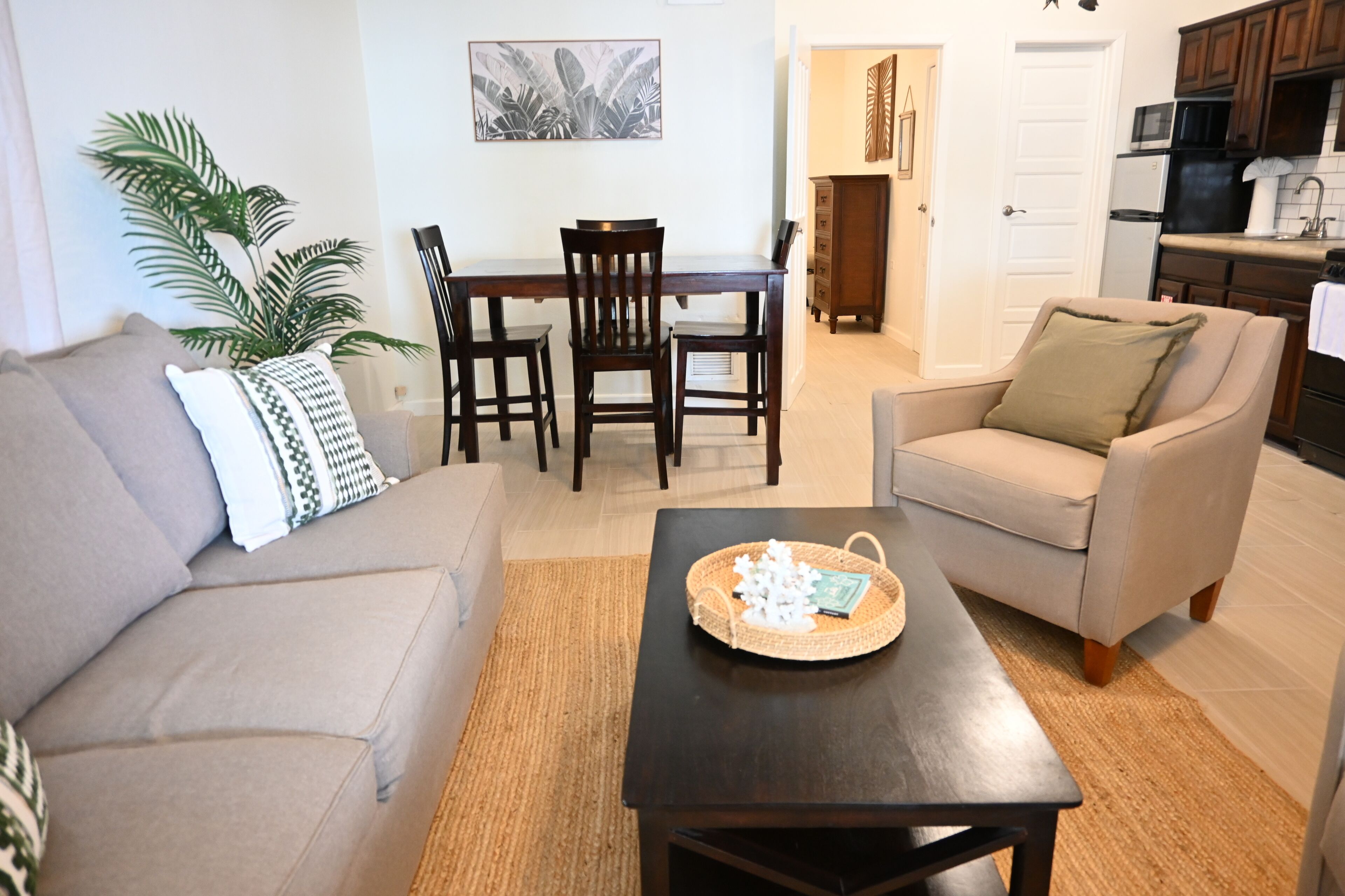 A living room with a sofa, armchair, coffee table, dining table with chairs, and a kitchen in the background.