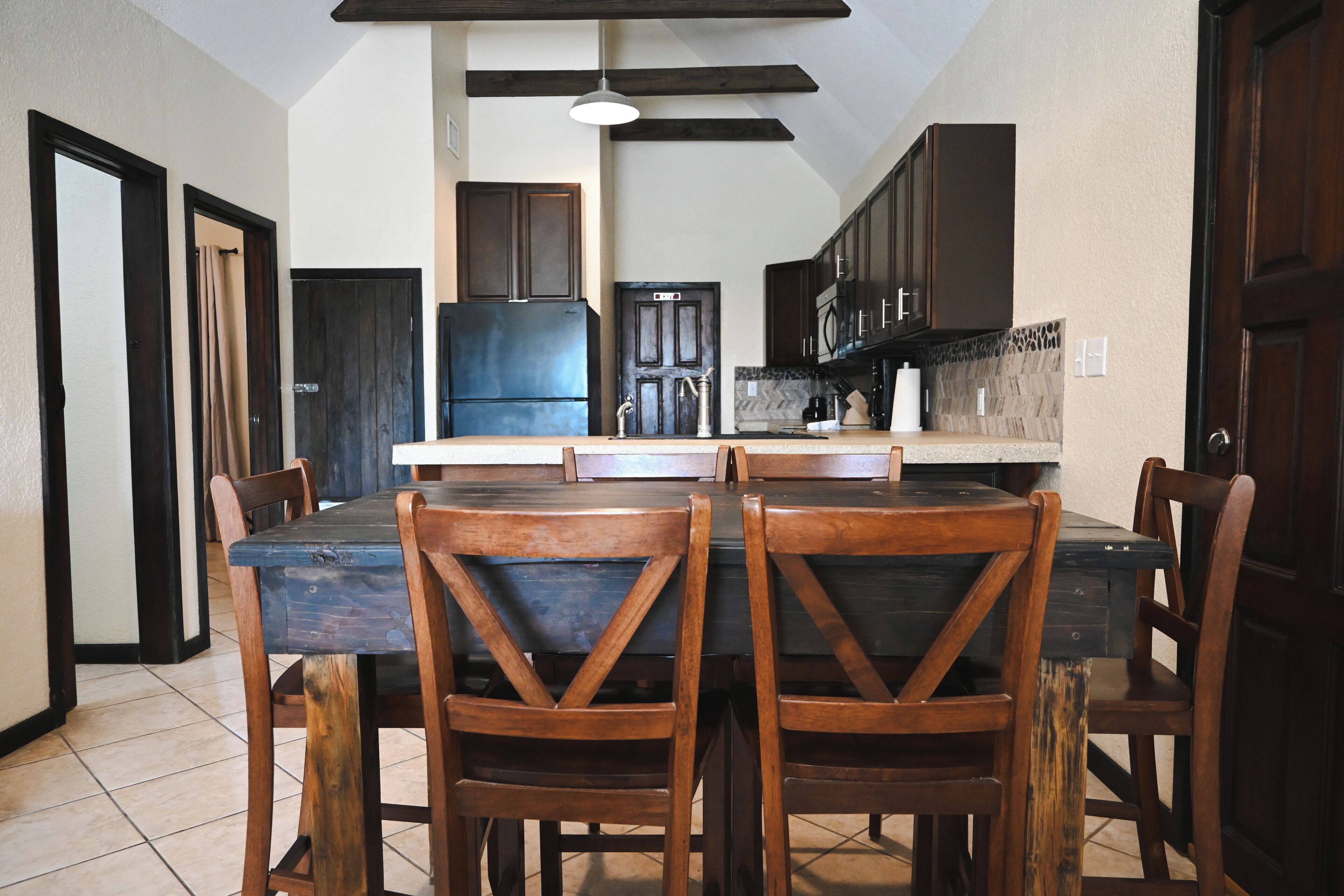 Un coin repas avec une table et des chaises en bois, une cuisine avec des meubles sombres et un réfrigérateur, et un sol carrelé.