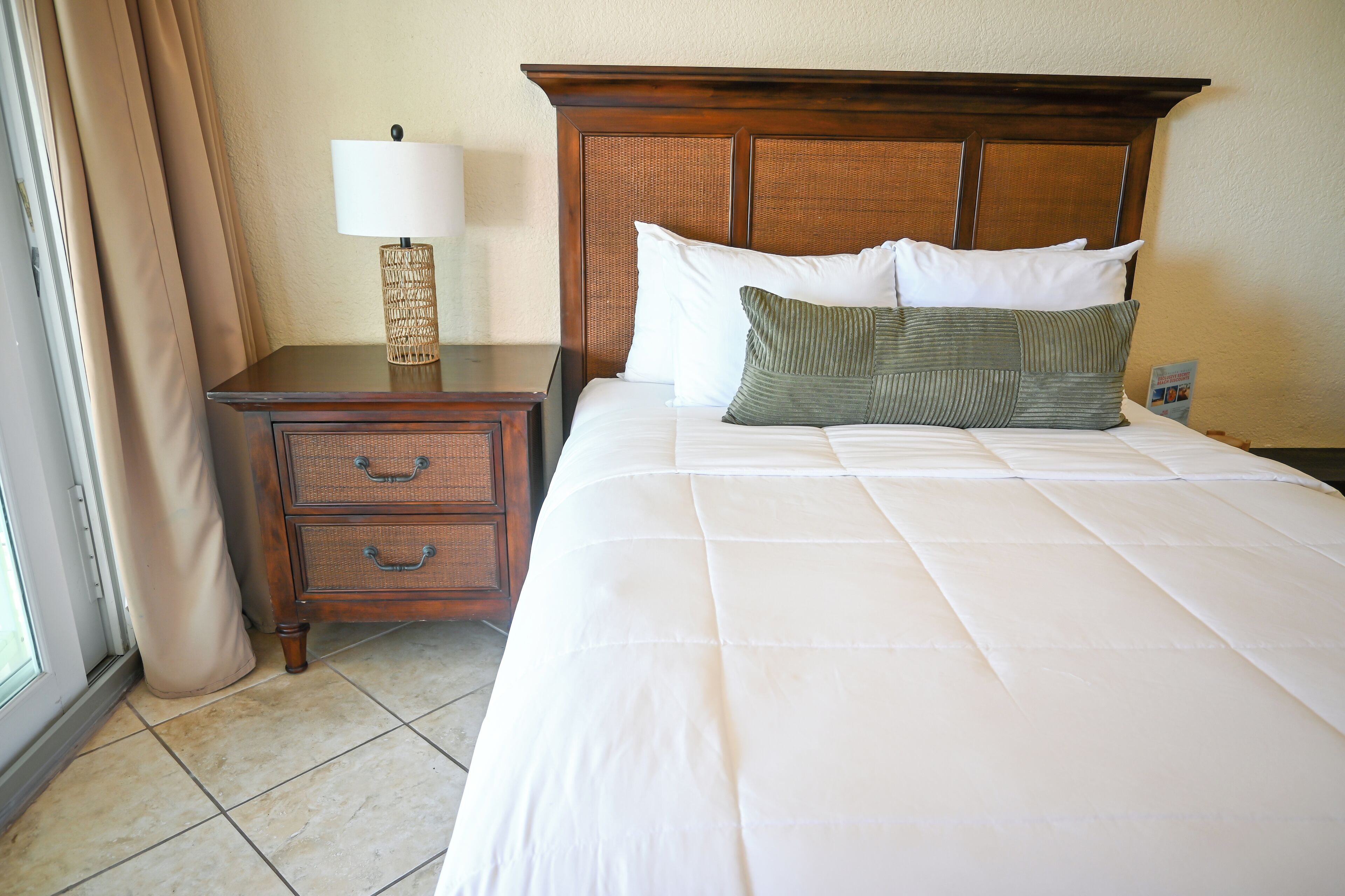 A neatly made bed with a wooden headboard, a bedside table with a lamp, and a small drawer unit.