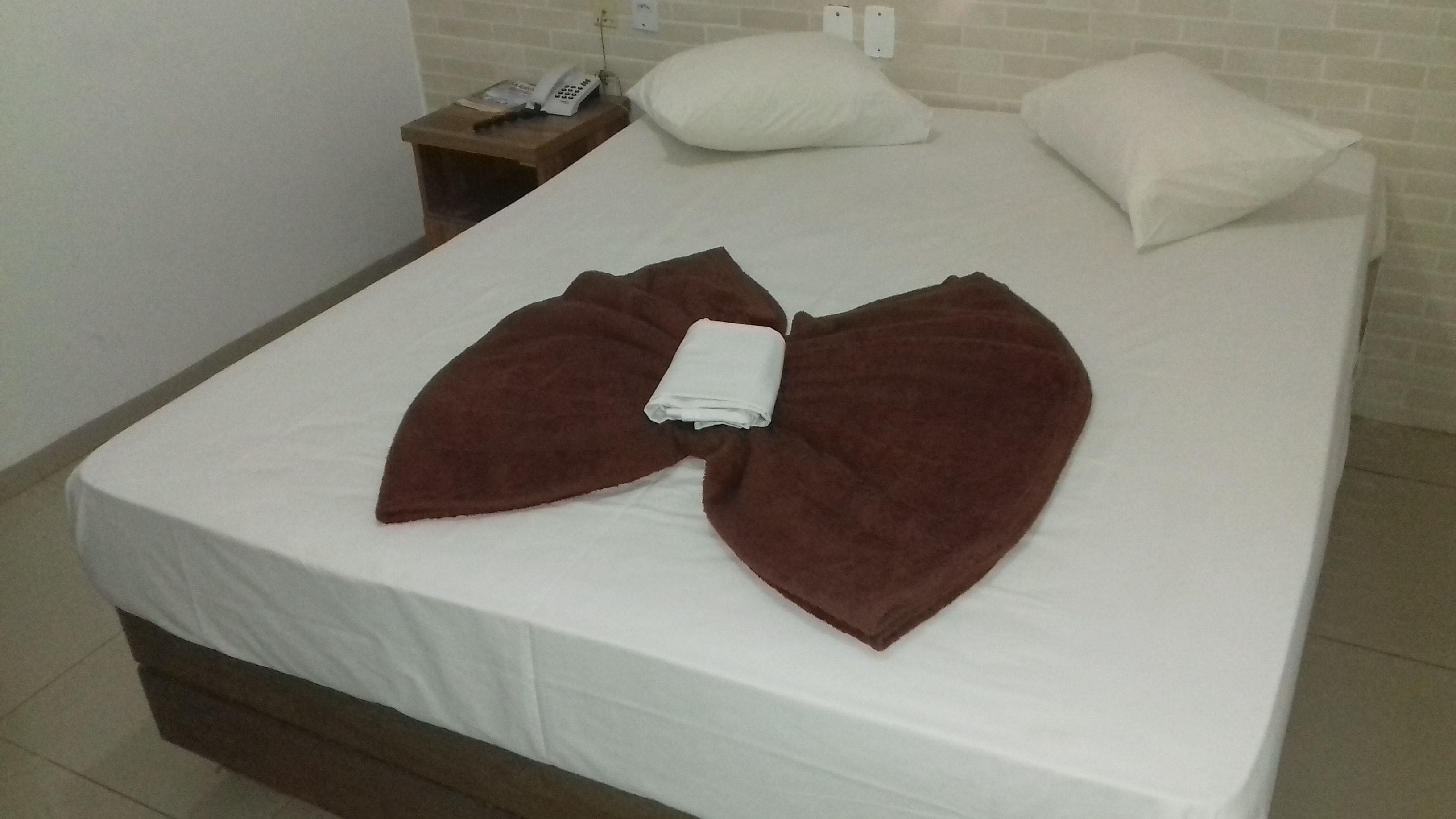 Een netjes opgemaakt bed met een gevouwen bruine handdoek in het midden, een wit kussen en een nachtkastje met een telefoon en een stopcontact.