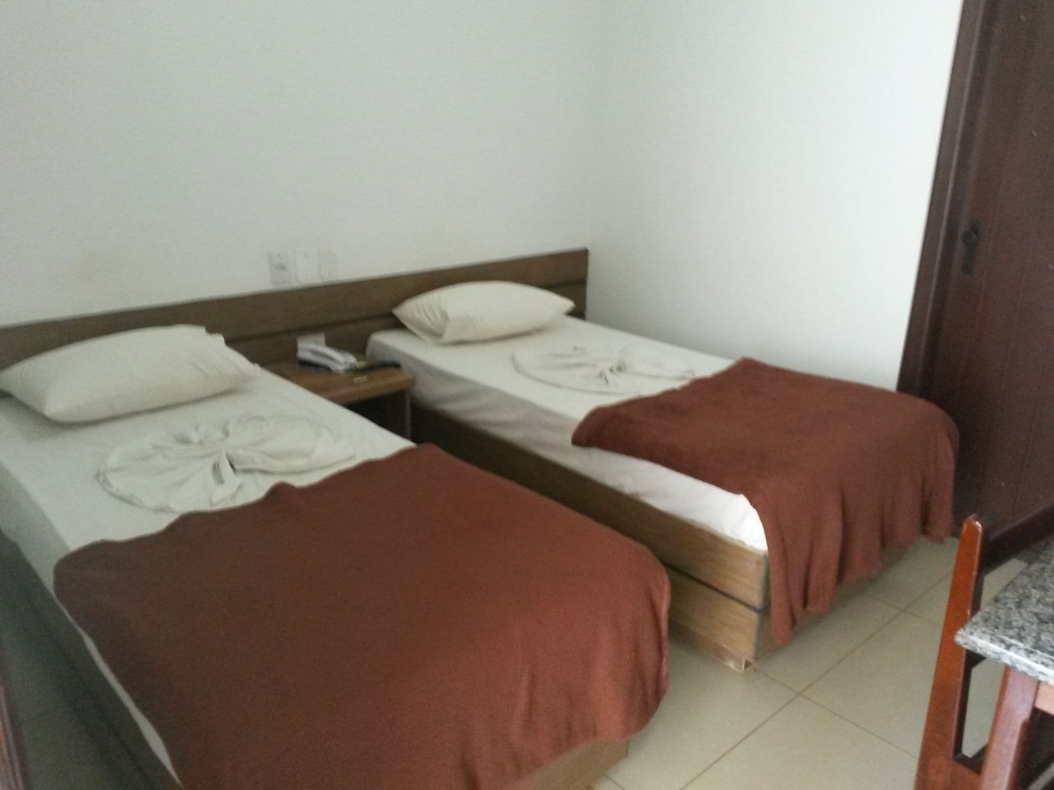 Una habitación con dos camas individuales, cada una con una almohada y una manta. Entre las camas, se encuentra una mesita de noche de madera con un teléfono y una lámpara. Se observa una silla de madera en la esquina.