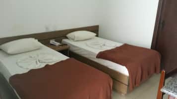 A room with two single beds, each with a pillow and a blanket. There is a wooden nightstand with a phone and a lamp between the beds. A wooden chair is visible in the corner.