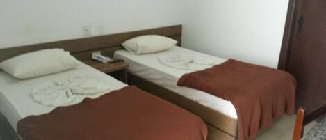Una habitación con dos camas individuales, cada una con una almohada y una manta. Entre las camas, se encuentra una mesita de noche de madera con un teléfono y una lámpara. Se observa una silla de madera en la esquina.