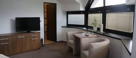 A hotel room with a bed, a TV on a wooden cabinet, a small table with a plant, two chairs, and a window with blinds.