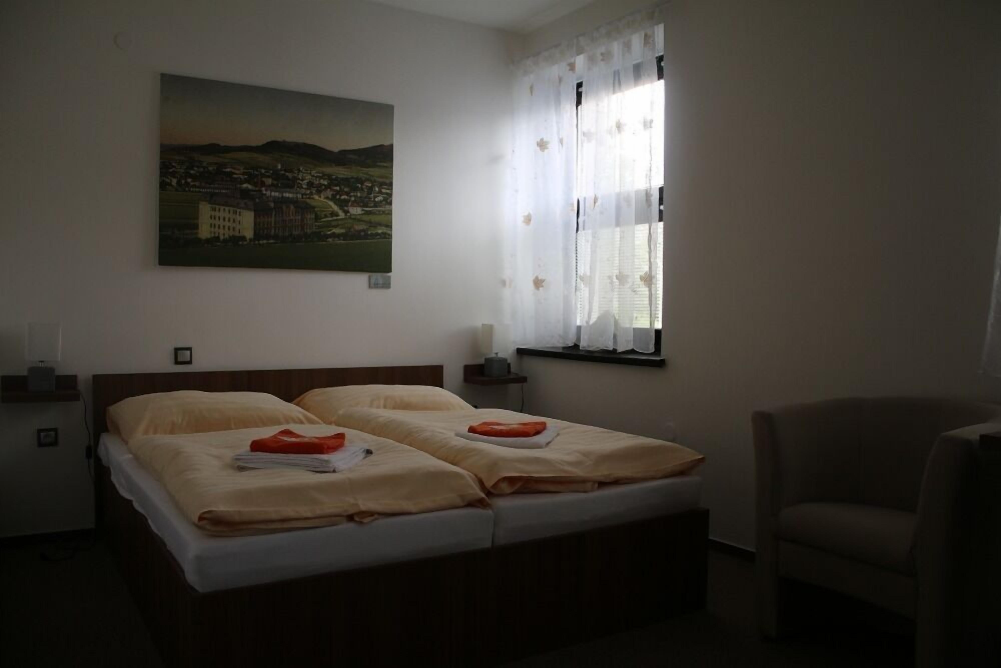 A double bed with beige bedding, a nightstand with a lamp, a chair, and a painting of a landscape on the wall.
