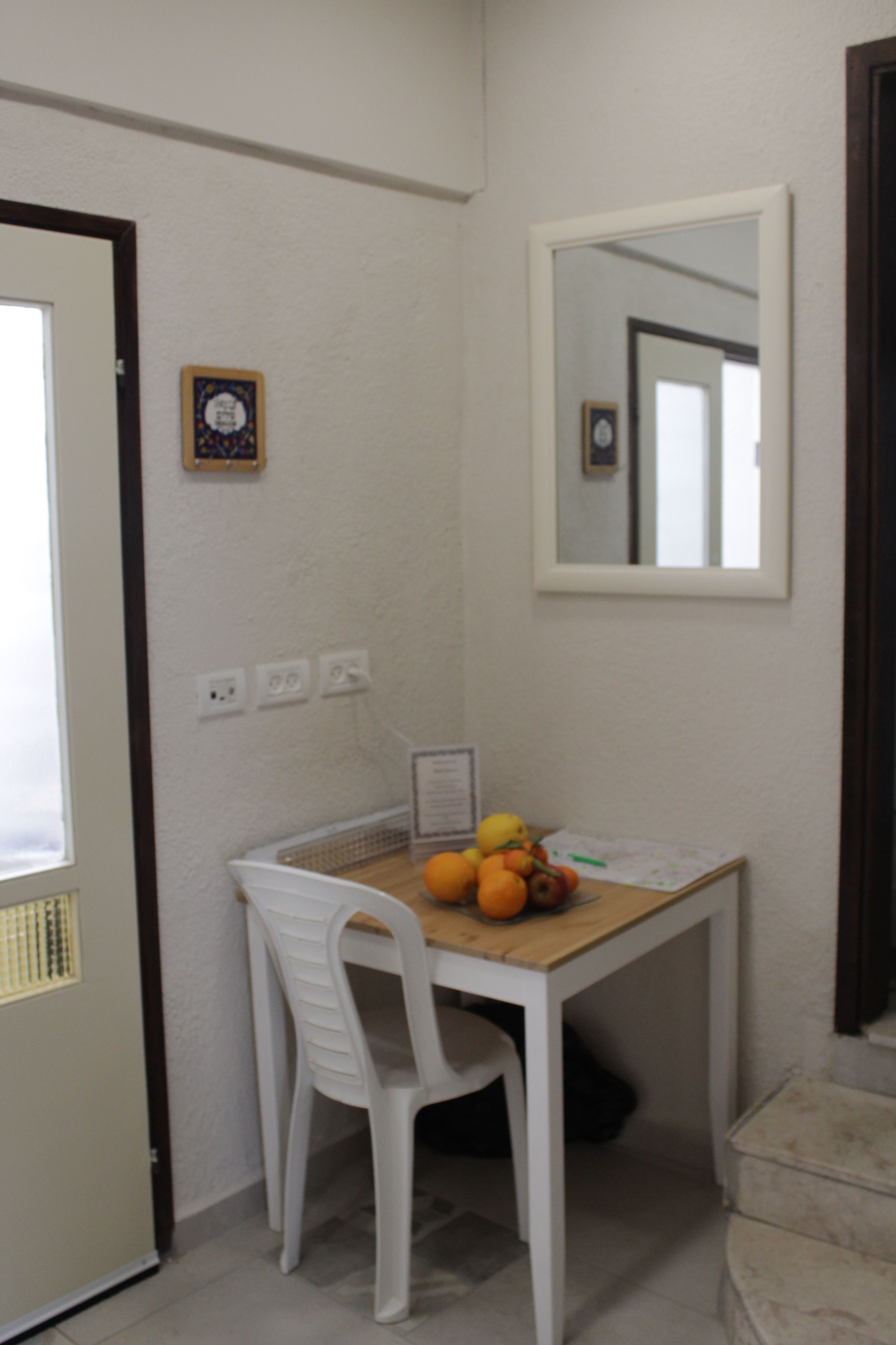 A small room with a wooden table, two white chairs, a mirror, and a fruit bowl with oranges and a banana.