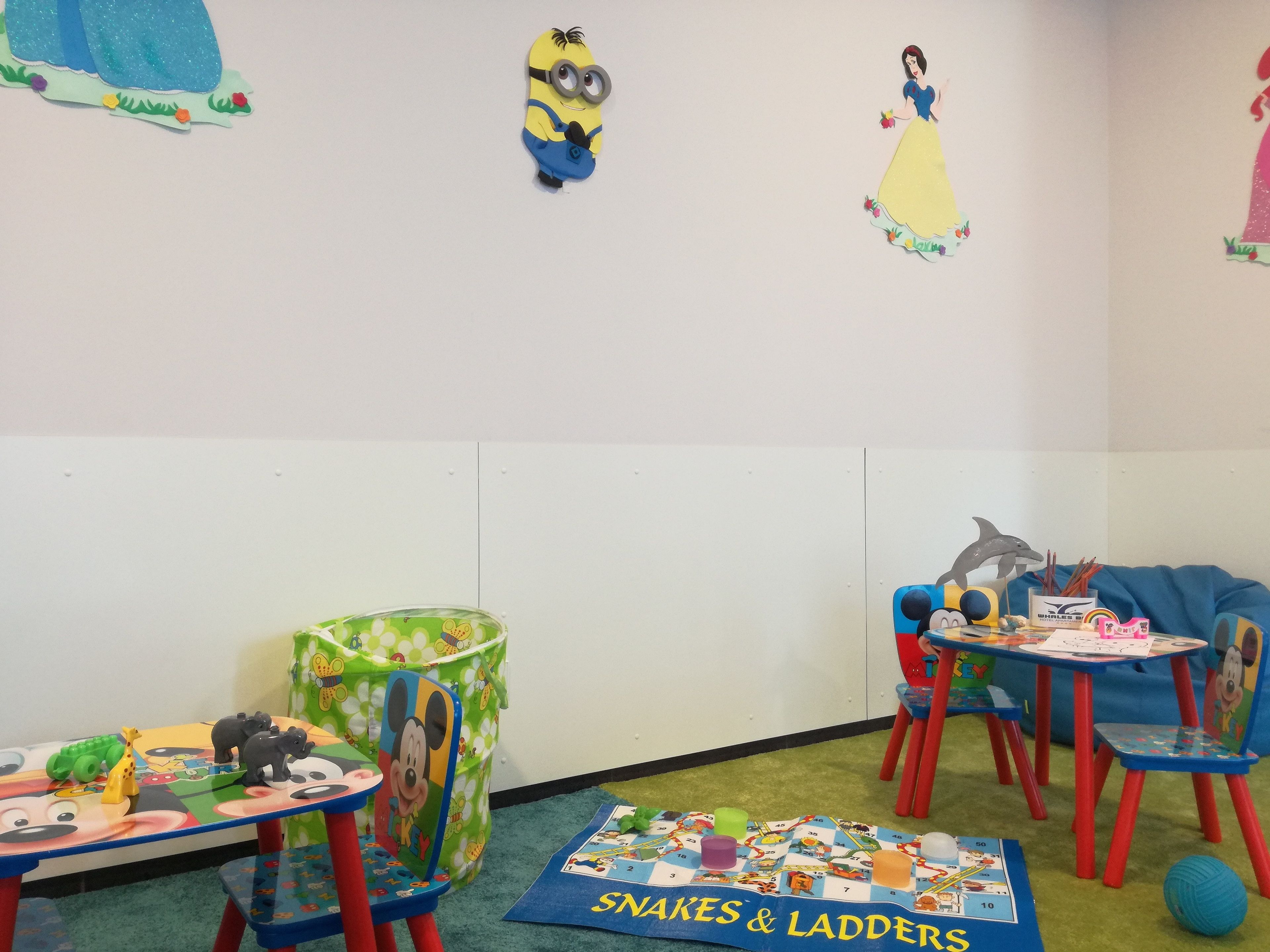 A children's playroom with colorful furniture, a play mat titled "Snakes & Ladders," and various toys.